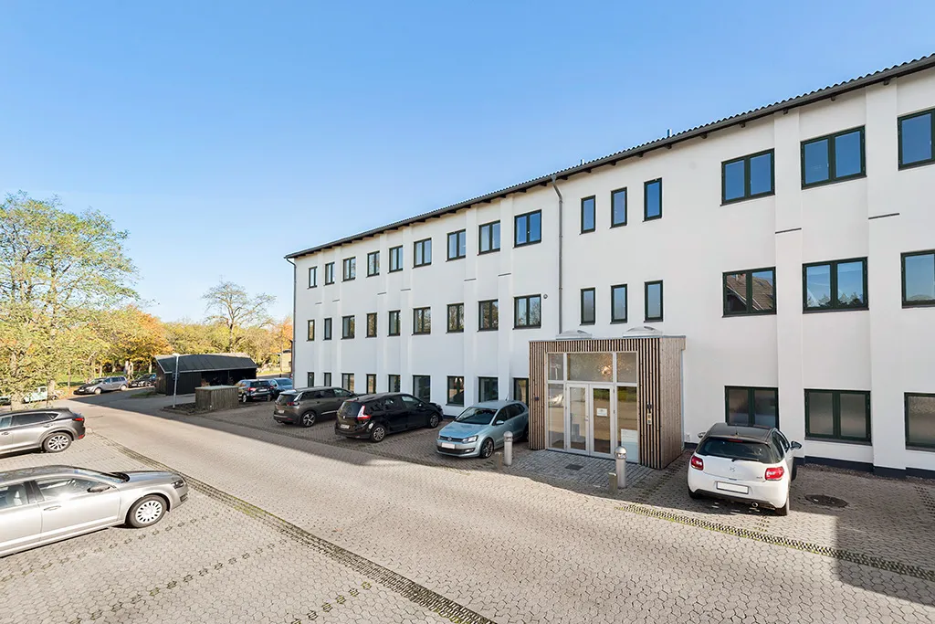 Hvidt bygningspark med parkeringsplads og udsigt til skov og parkerede biler under klar himmel