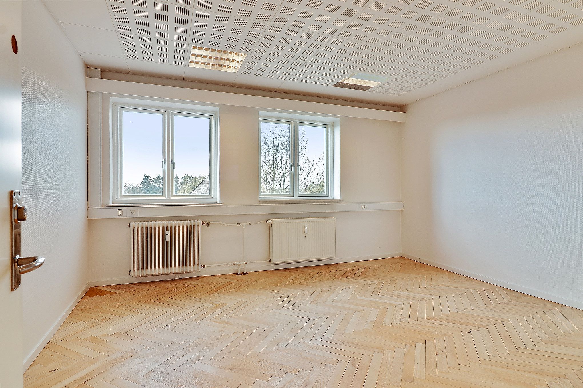 En tom rum med lyse trægulve og to store vinduer på en væg samt to radiatorer og hvide vægge