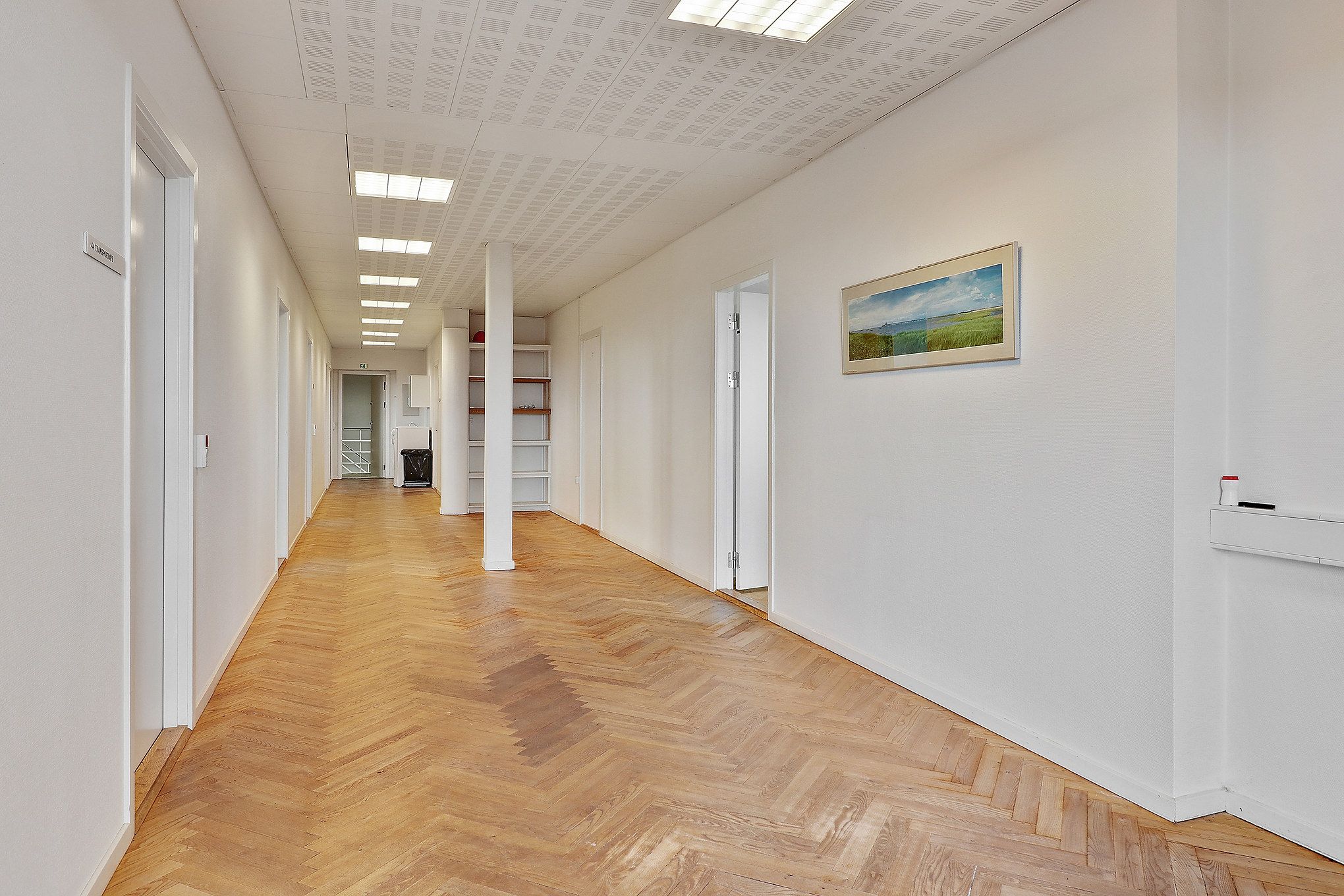 Kort korridor med lysegrå vægge og parketgulv samt kunstværk og åbne døre i baggrunden