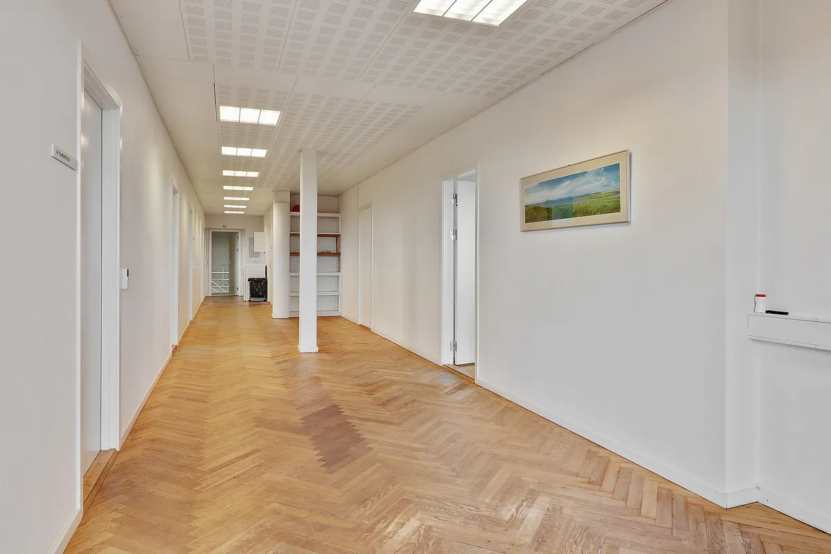 Kort korridor med lysegrå vægge og parketgulv samt kunstværk og åbne døre i baggrunden