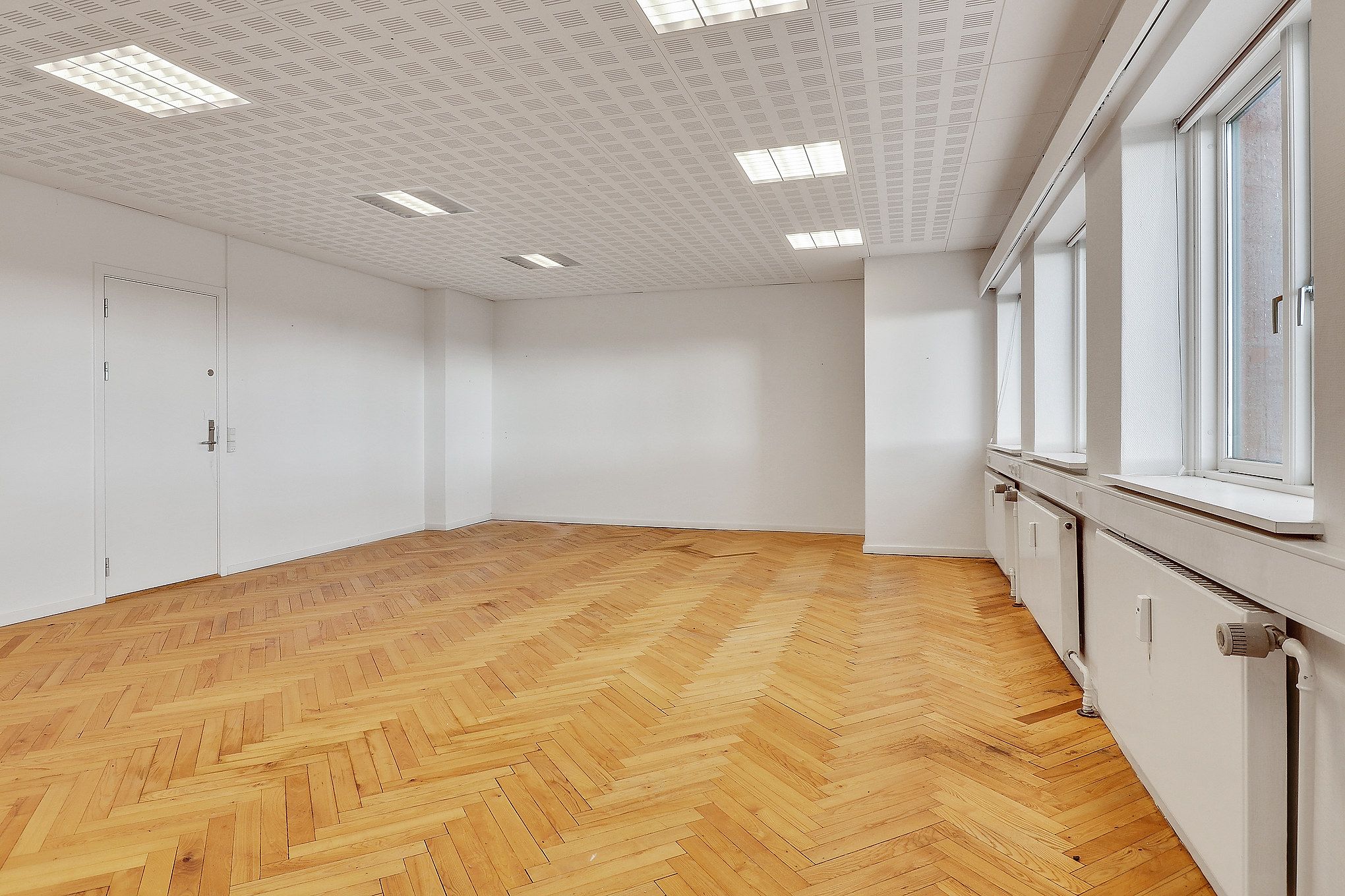 Et stort, tomt rum med lysegult trægulv og hvide vægge samt vinduer og døre. Der er fire radiatorer og tre store vinduer med