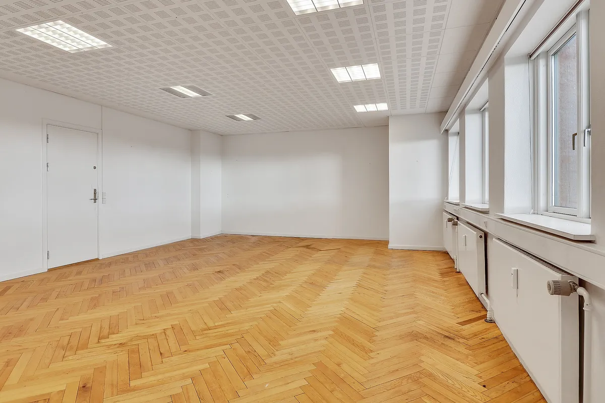 Et stort, tomt rum med lysegult trægulv og hvide vægge samt vinduer og døre. Der er fire radiatorer og tre store vinduer med