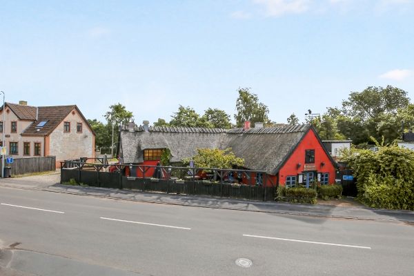 Rød kro med lyseblå ruder og tag dækket af halm står på hjørnet af en tomte med grønne buske og træer