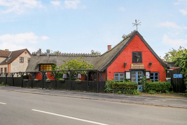 En gammel rød stue på Nyvej 18 i Liseleje med tegltag, vinduer og lukket dør. Butikken har en søjle og vinduer på siderne.