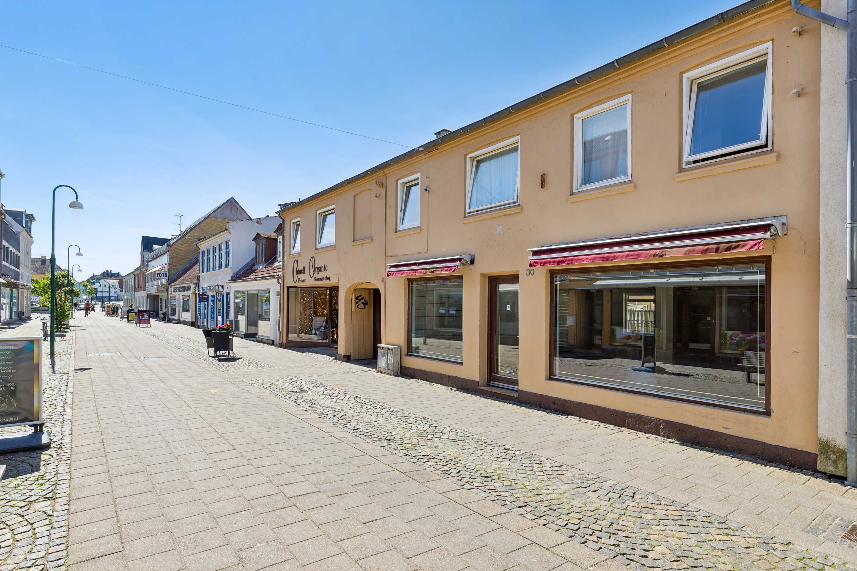 En tom gågade med forretningslokaler og gåbord. Lys dag med lyseregn og sol. En søjle med gadelampe og en annonceskil.