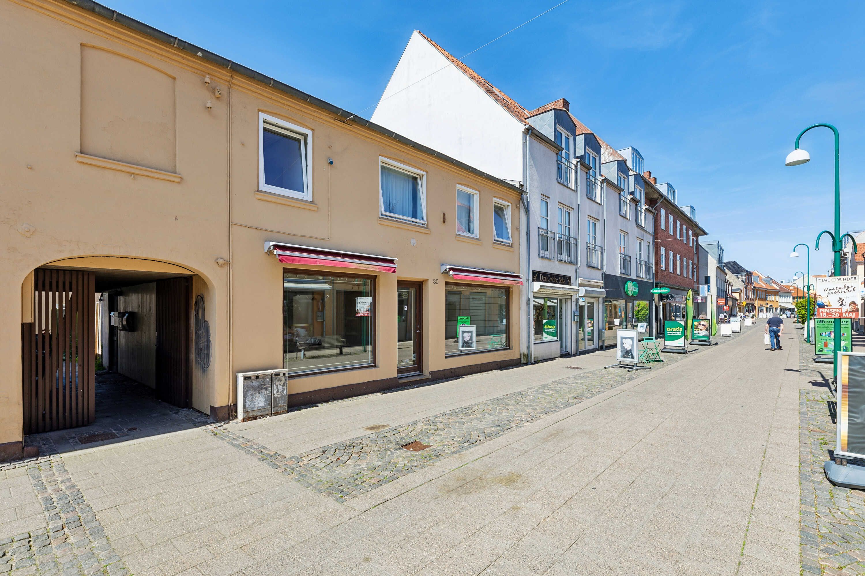 En tom gågade med små butikker og reklameskilte, der vender mod højre og en person med køb i hånden i baggrunden