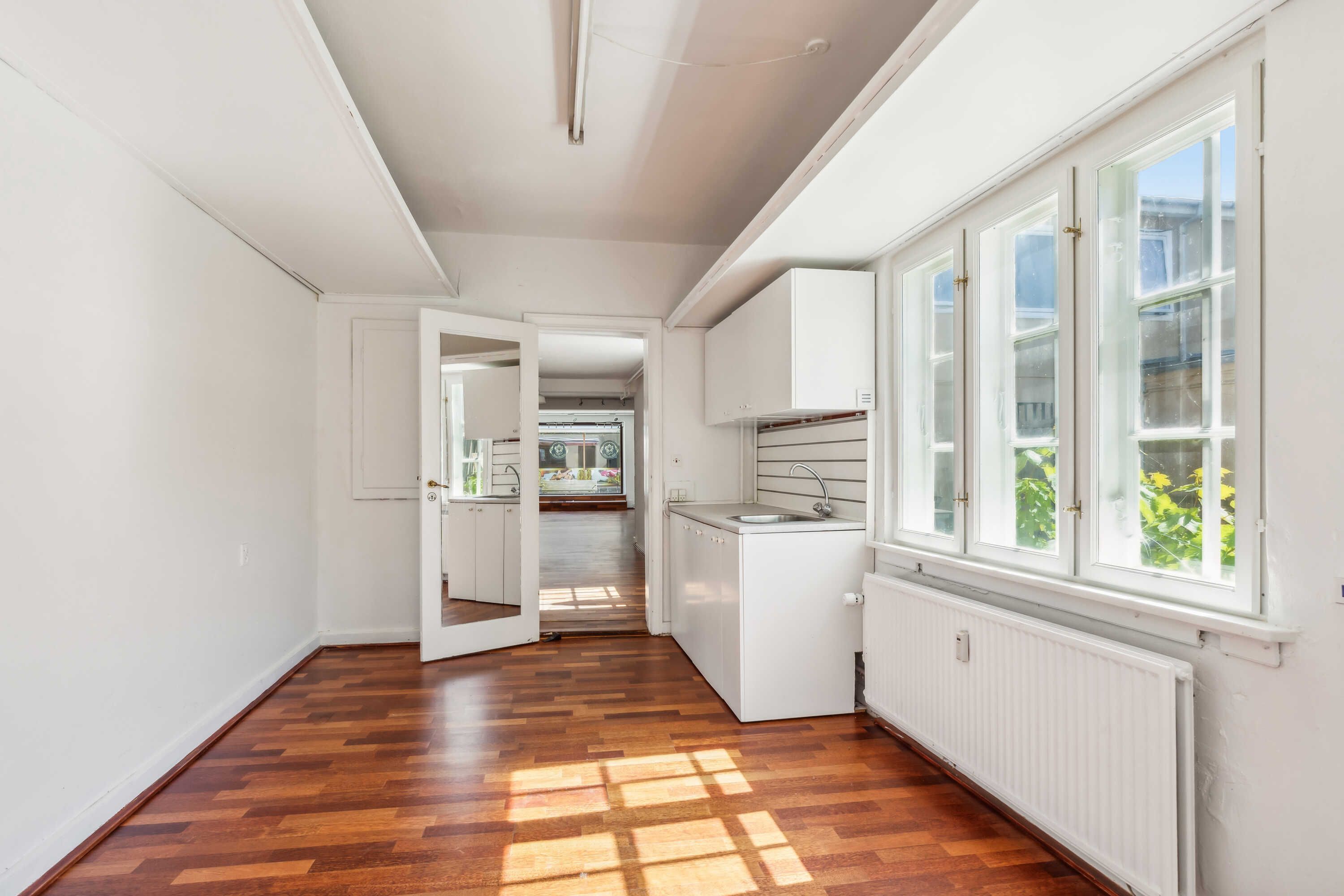 En tom lejlighed med hvide vægge og trægulv og et køkken med hvide skabsvægge og en radiator under vinduet