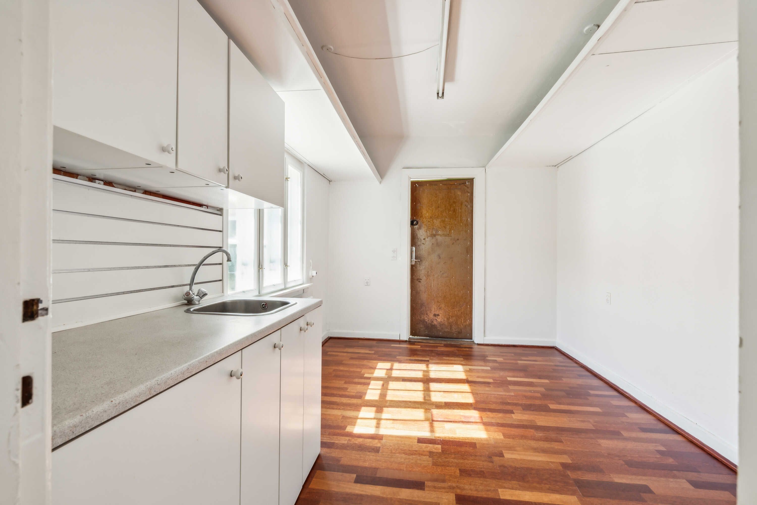 En minimalistisk køkken med hvide vægge, en rusten dør, og et moderne vask med en sølvfarvet vandhane