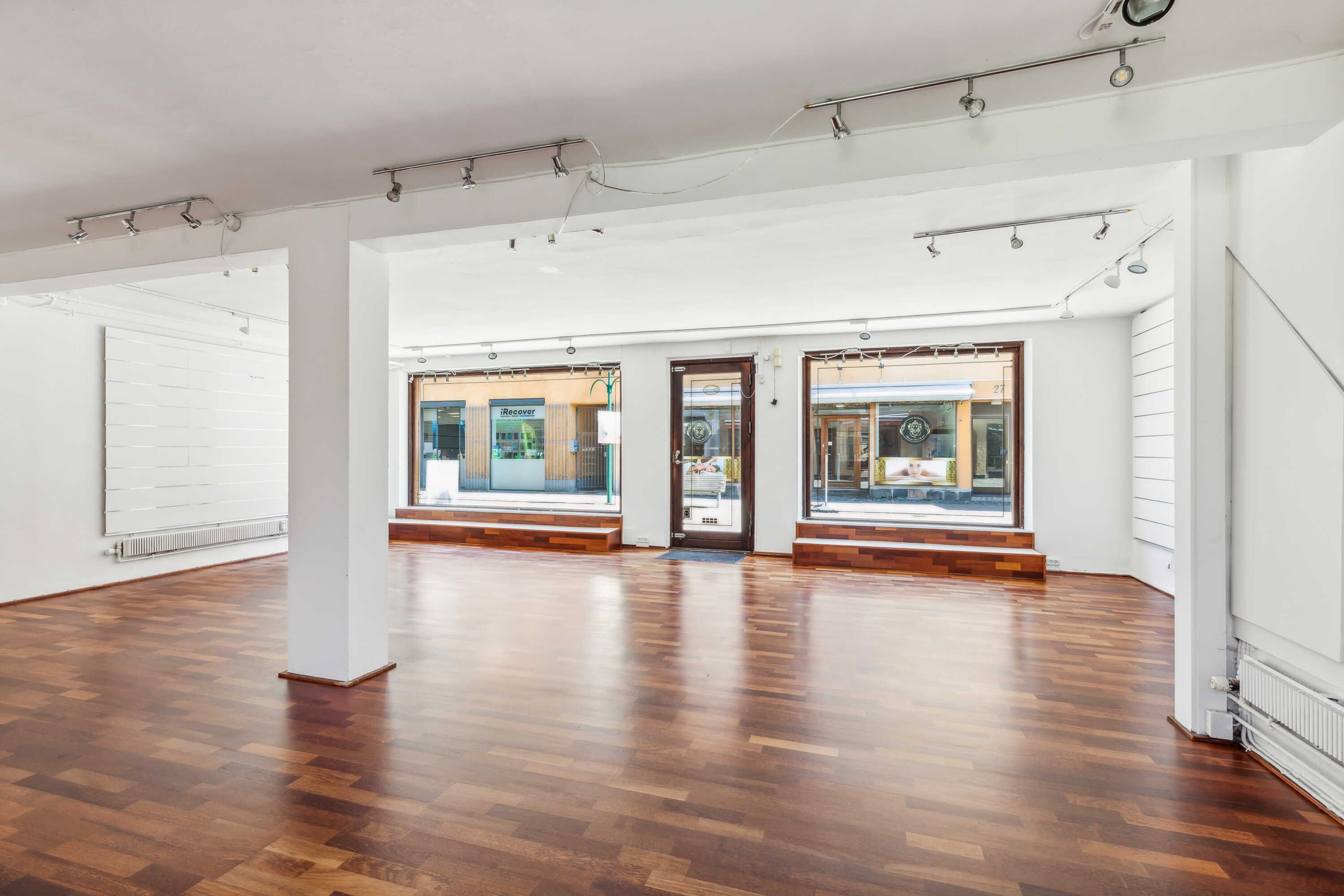 Tom rum med hårdt trægulv og hvide vægge, store vinduer med glasdøre, spor af indvendige radiatorer og lysekroner i loftet