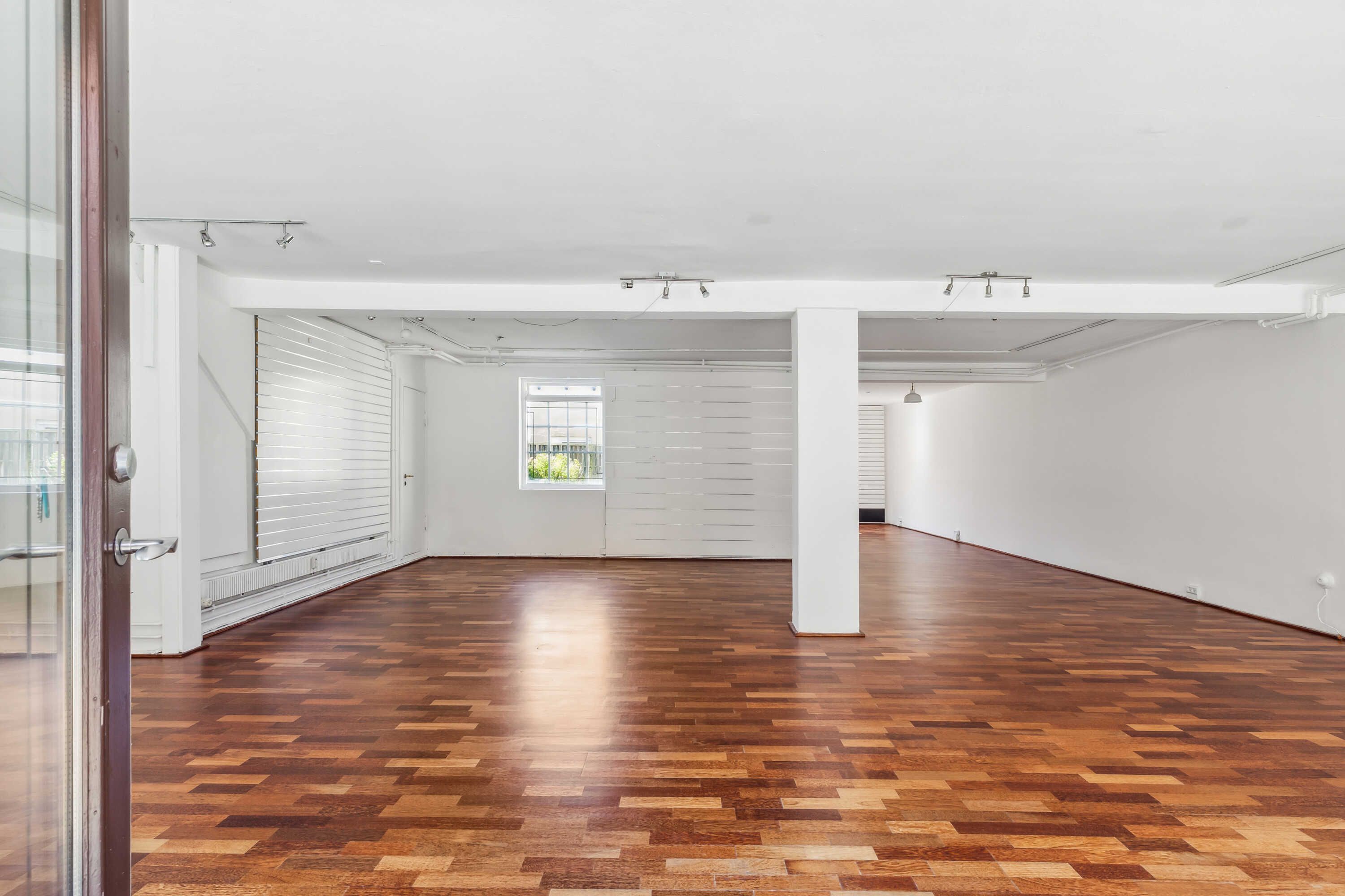 En stor, tom lejlighed med trægulve og hvide vægge og loft. Væggene er delvist dækket af hvide skodder og der er to små