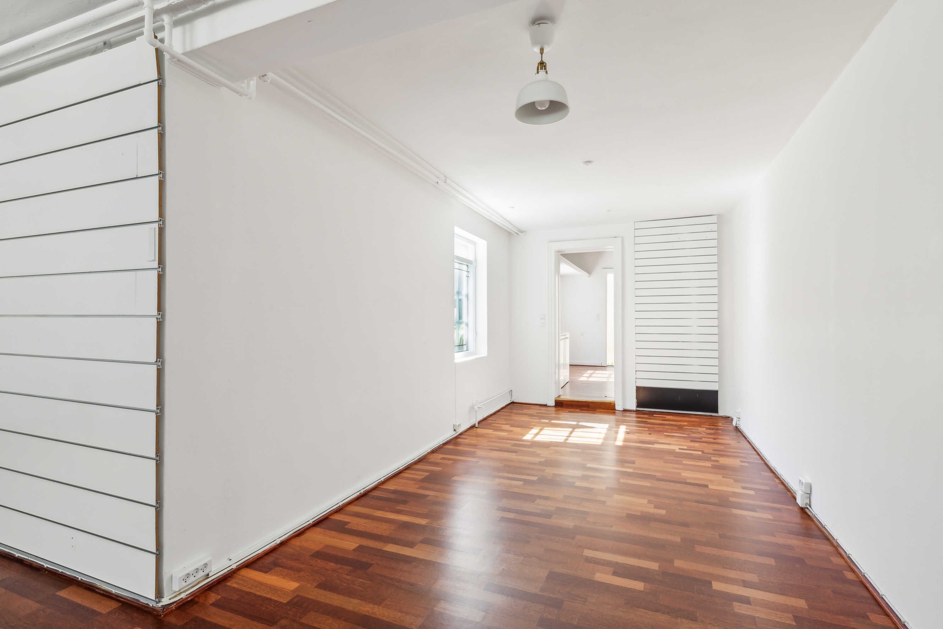 Et tomt rum med hvide vægge og trægulv samt et lysekron og et vindue på højre side