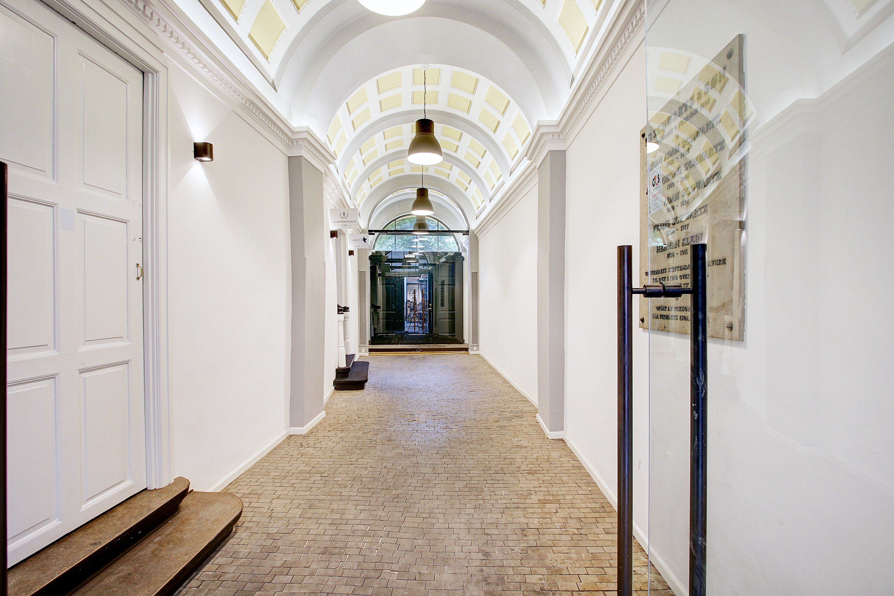 Hal med hvide vægge, lysekroner, mønstret loft og lysekroner. Indkørselsdør med skrift på den og møbler i midten.