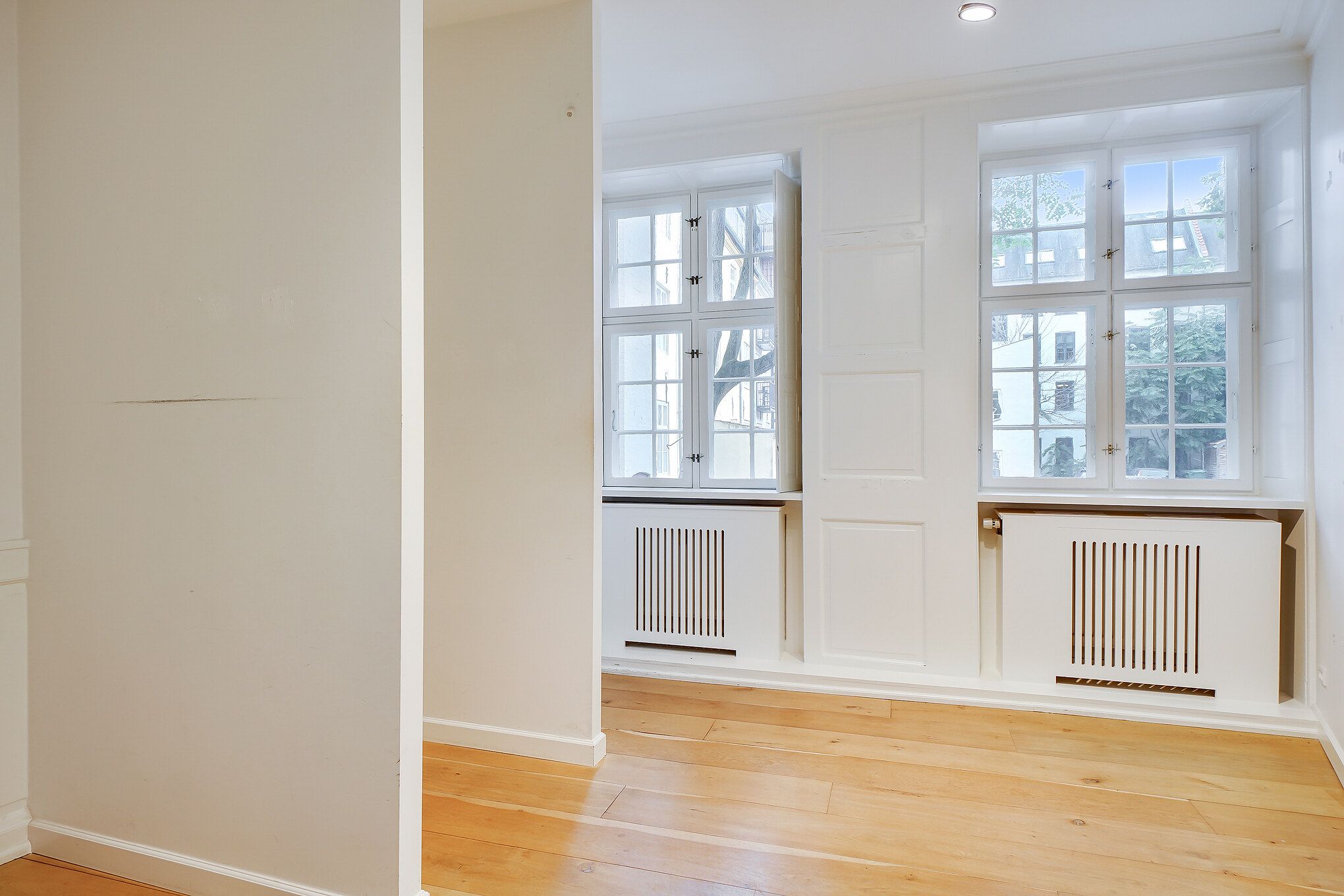 Værelse med hvide vægge og hårdt trægulv, to store vinduer med hvide rammer og radiatorer nederst i hvide.