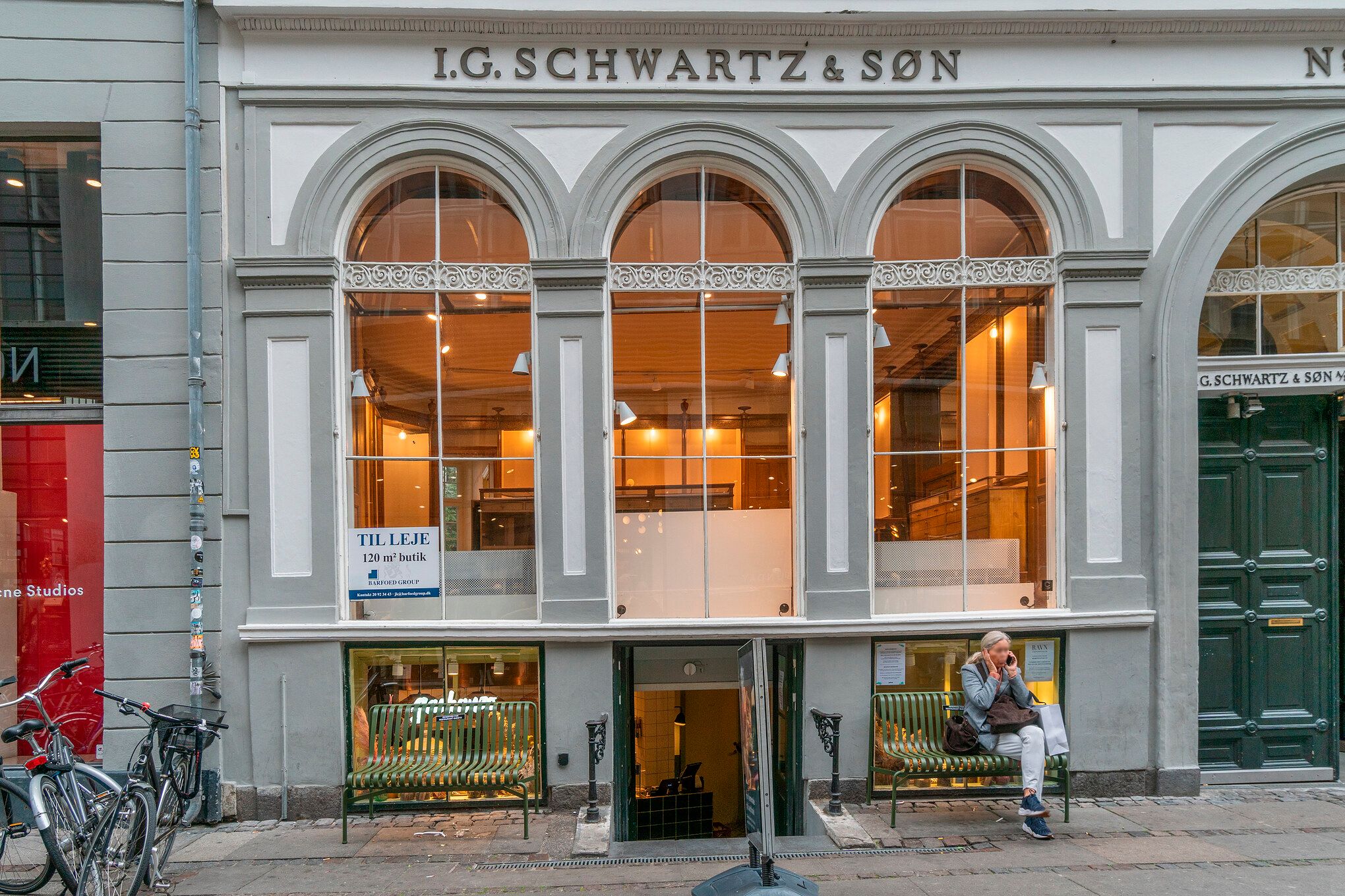 En dame sidder på en grøn bænk og snakker på mobiltelefonen uden for indgangen til L.G. Schwartz & Son