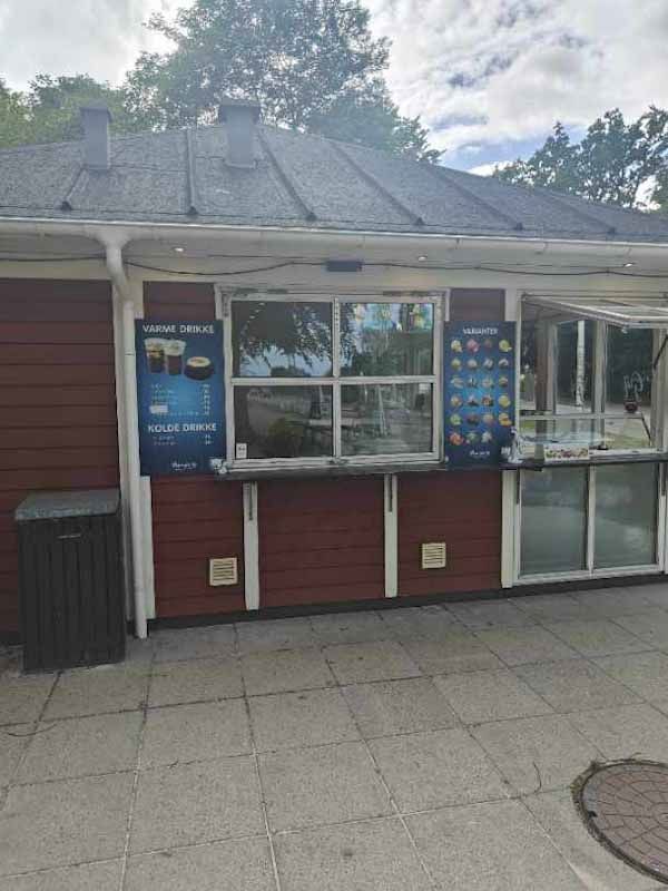 En rødmalet bygning med glasvinduer og skilte med tekst og billeder er i baggrunden, og en skraldkur er i forgrunden