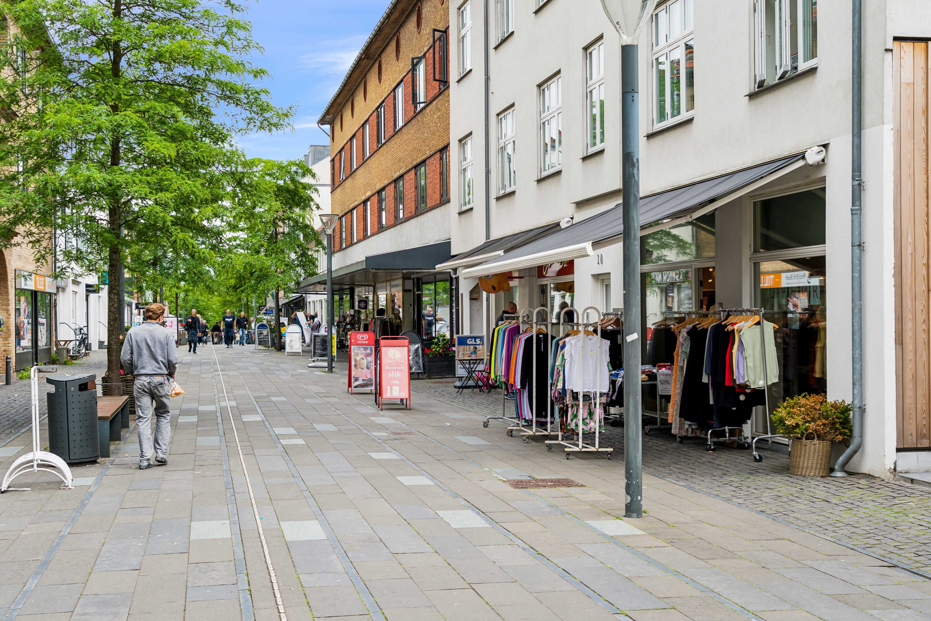 En mand går på en fredelig gågade med mange butikker, træer og blomsterpotter langs vejen, med en buskede plante og