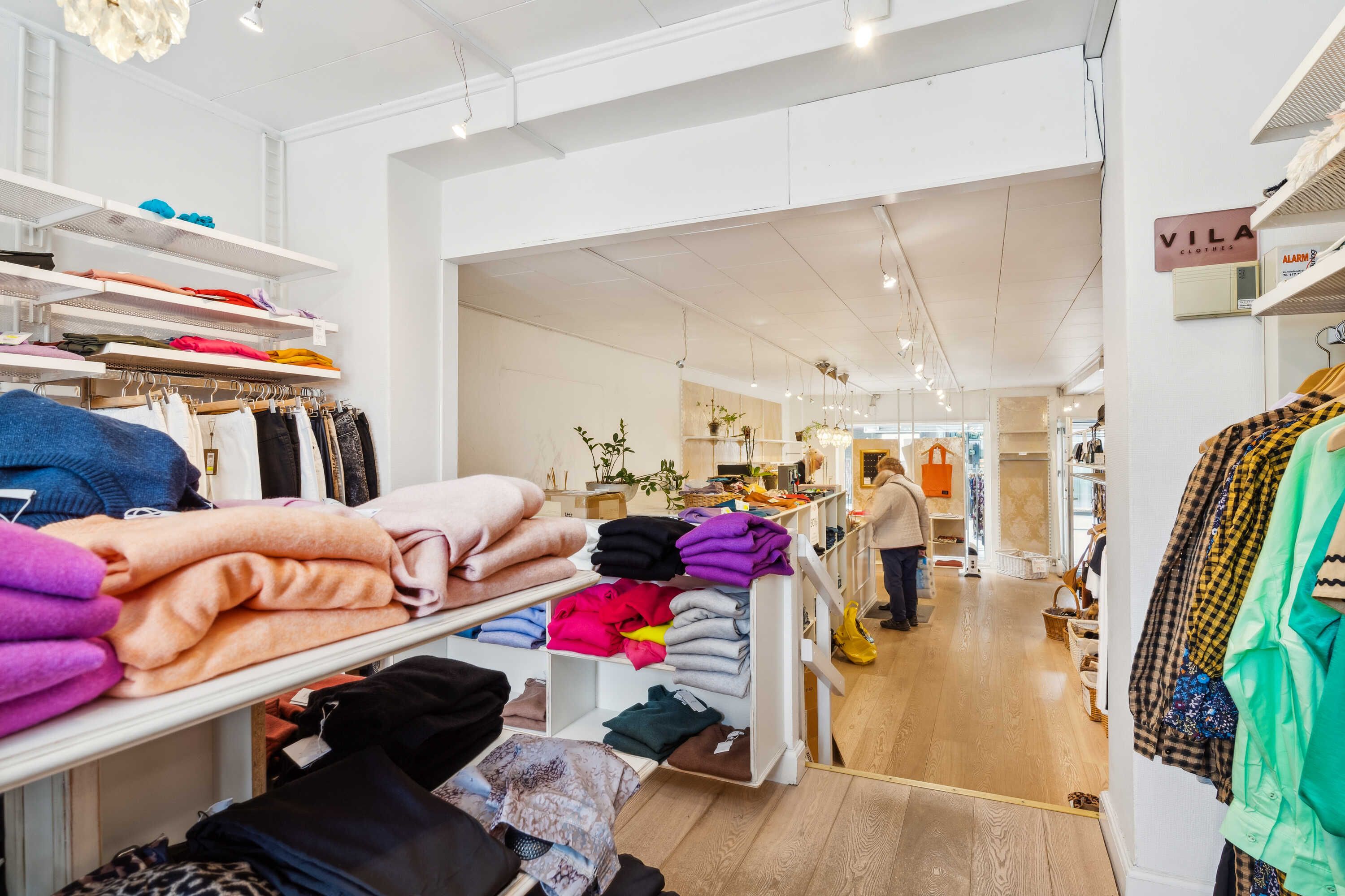 Forretningslokal med tøj på hylder og gulv, hvidt loft, lysekroner og trægulv
