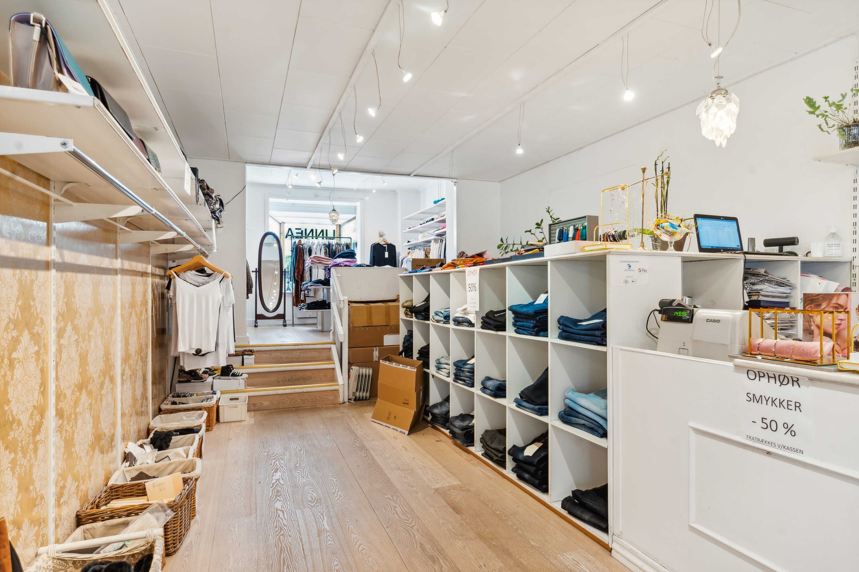 Moderne, velorganiseret tøjforretning med lyse gulve og hylder, udsalgstavle, lysekroner og plante.