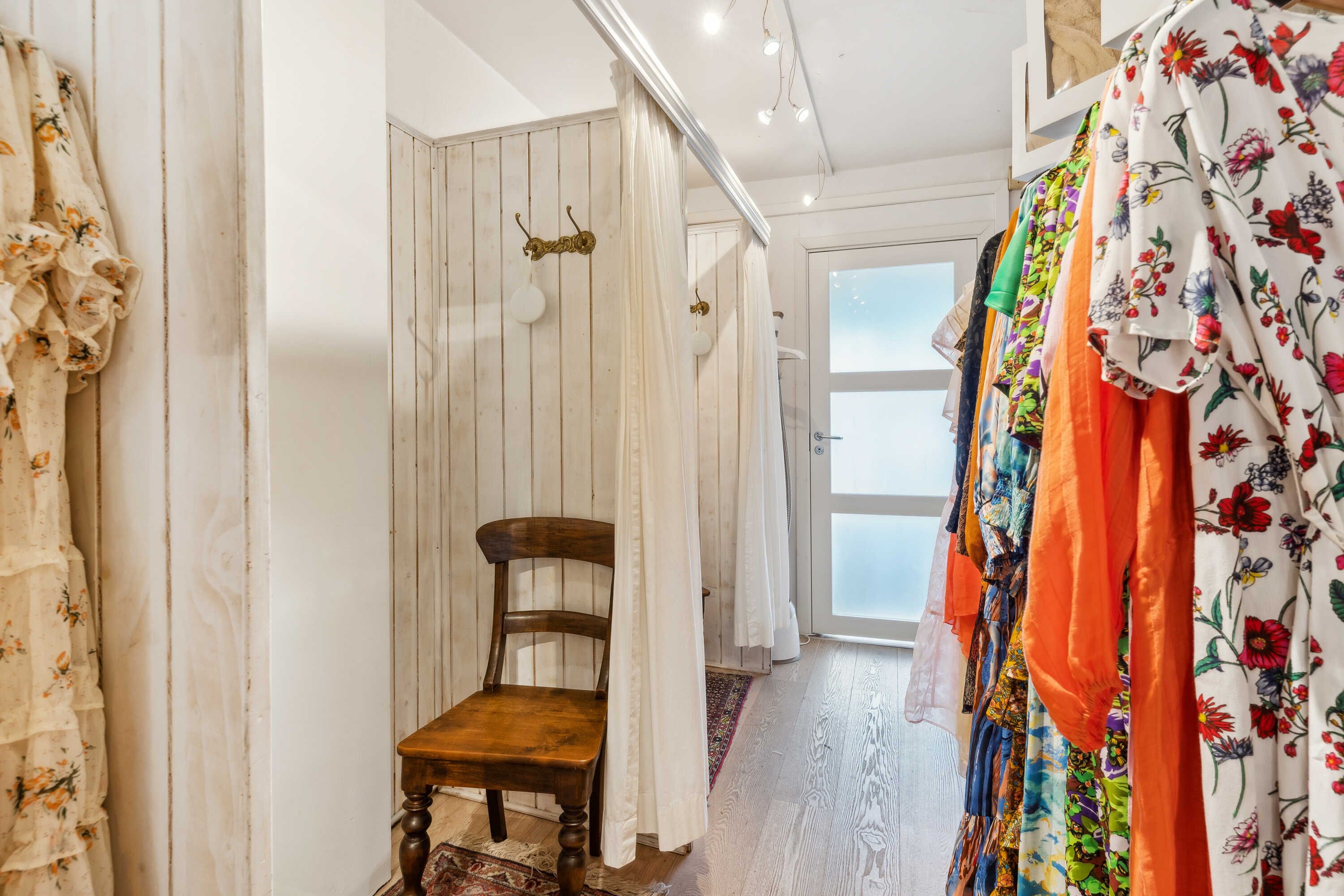 En lille indgangspassage med en træstol, lysekroner og tøjophængere med tøj i farverige mønstre og blomsterprint