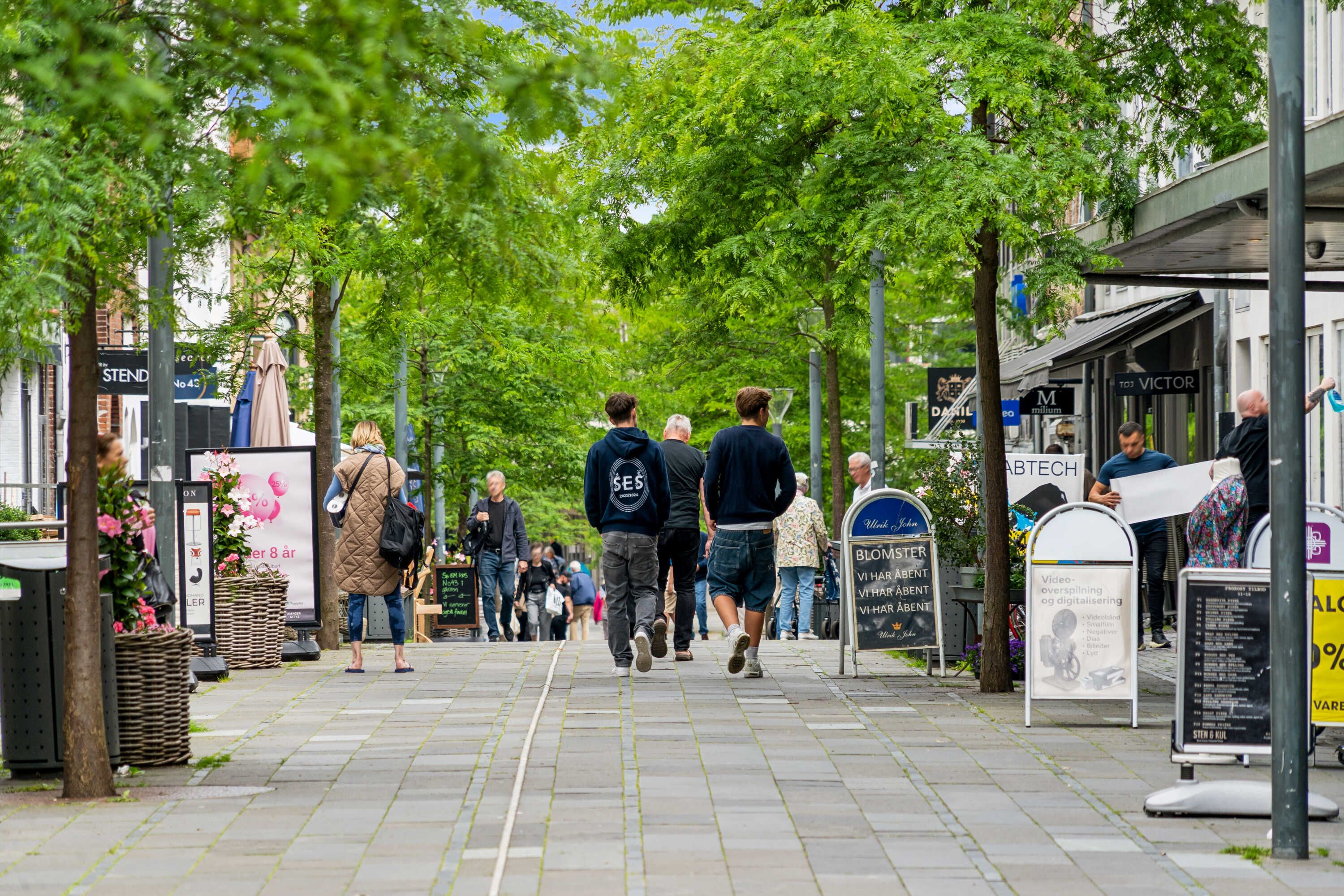 En gågade i Hørsholm med folk, gamle bygninger, butikker og grønne træer, som giver skygge i solen