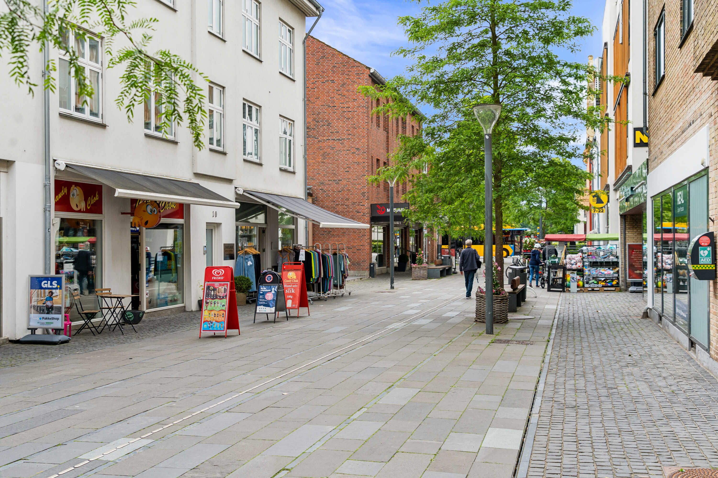 Butikslokale med aftenaviser og tøj på gaden, folk og gågade med gamle bygninger og grønne træer, og skilte med AED og N.