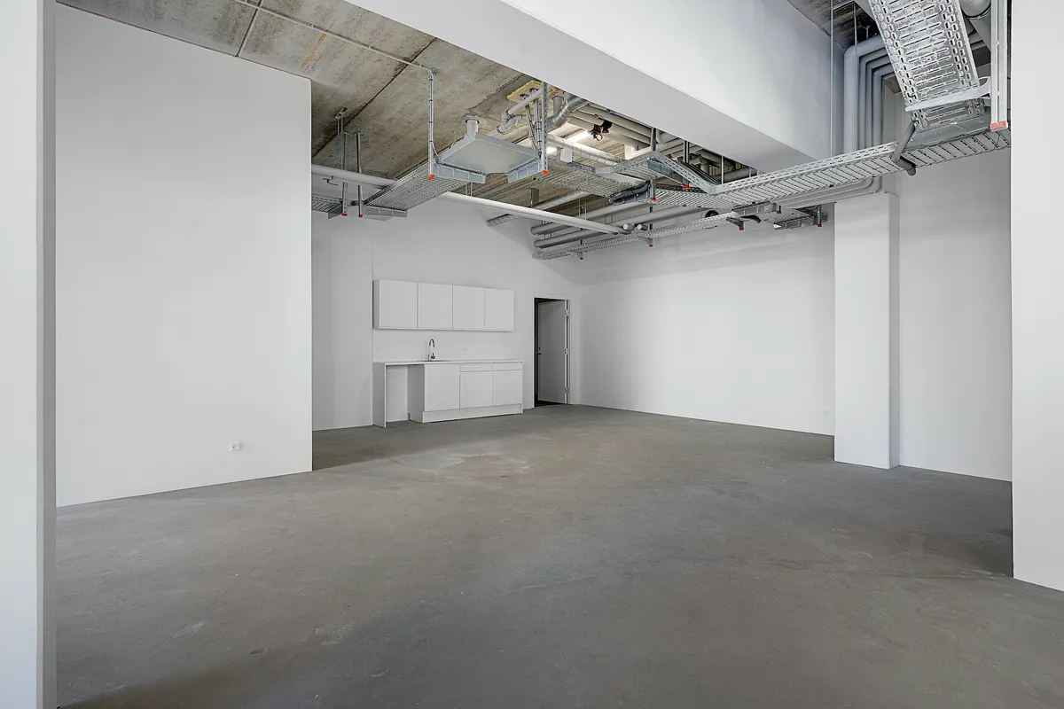 En stor og hvid stue med lysegul gulvbelægning, hvide vægge og lysekroner samt højtliggende rør og kabelkanaler