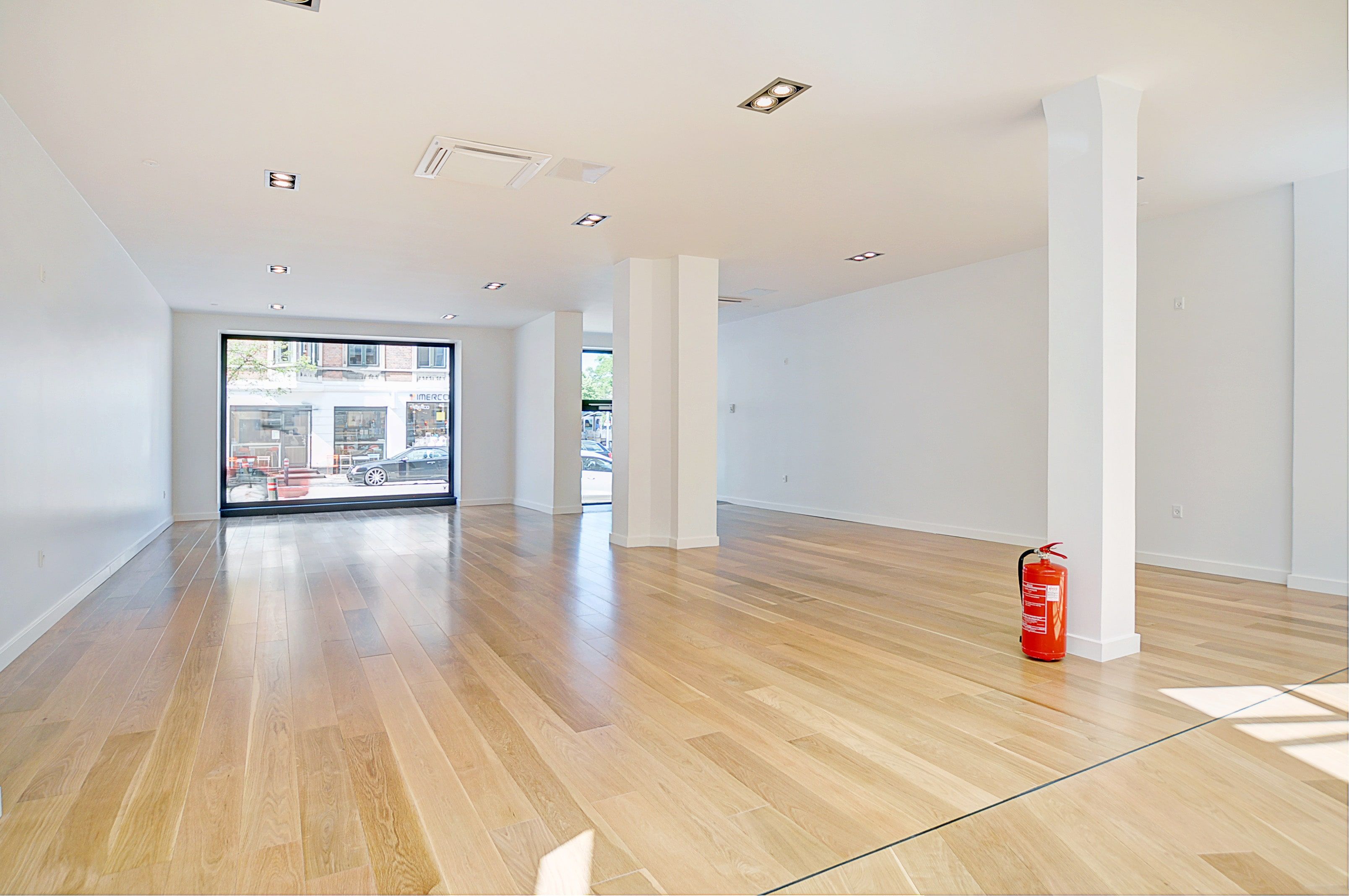 Store lyse lokaler med lysegul gulvbelægning og hvide vægge samt gulvvarme og store vinduer. Et rød brandværn til højre og