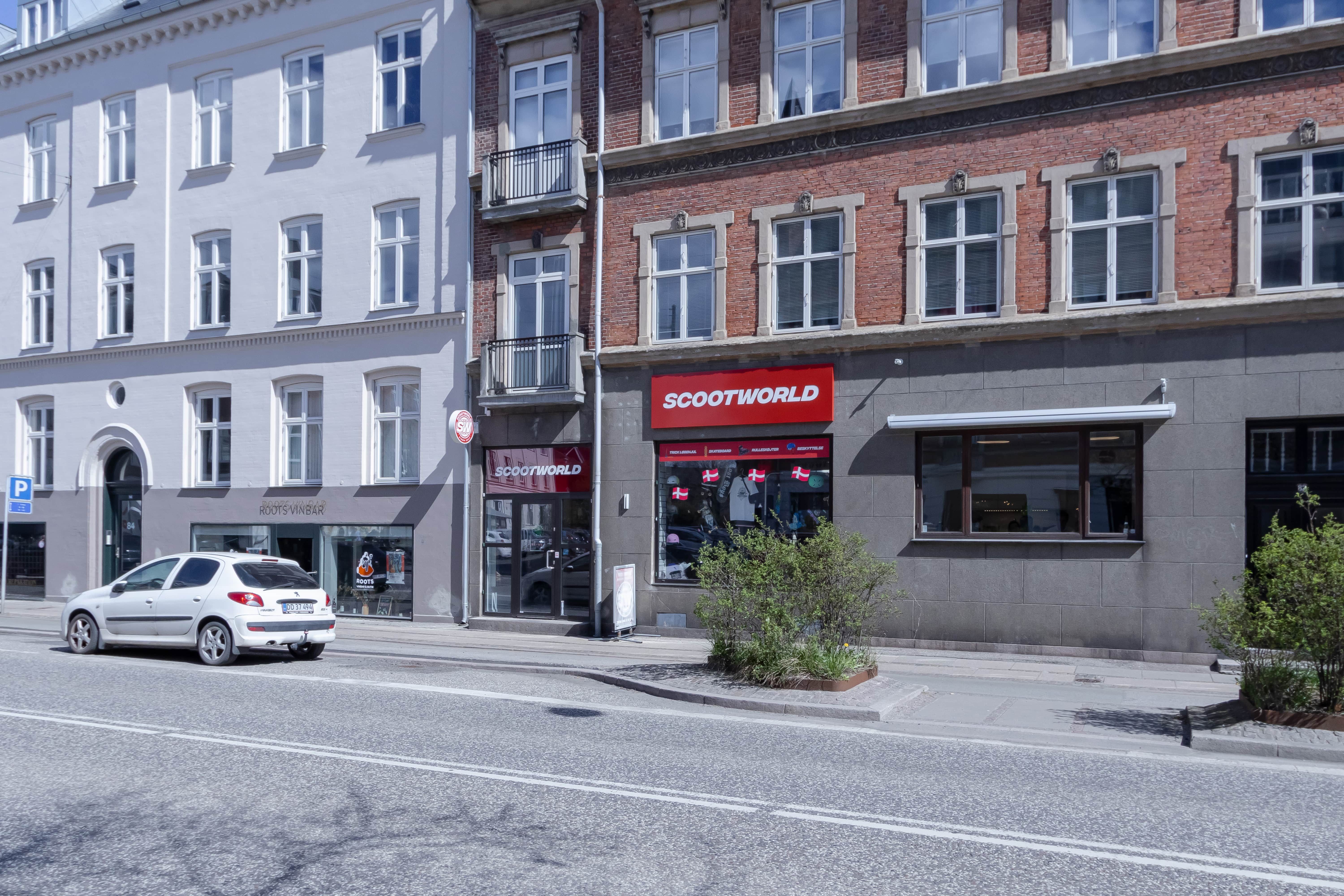 En Peugeot bil kører på gade med et butiksvindue med teksten Scootworld og en bevoksning i forgrunden