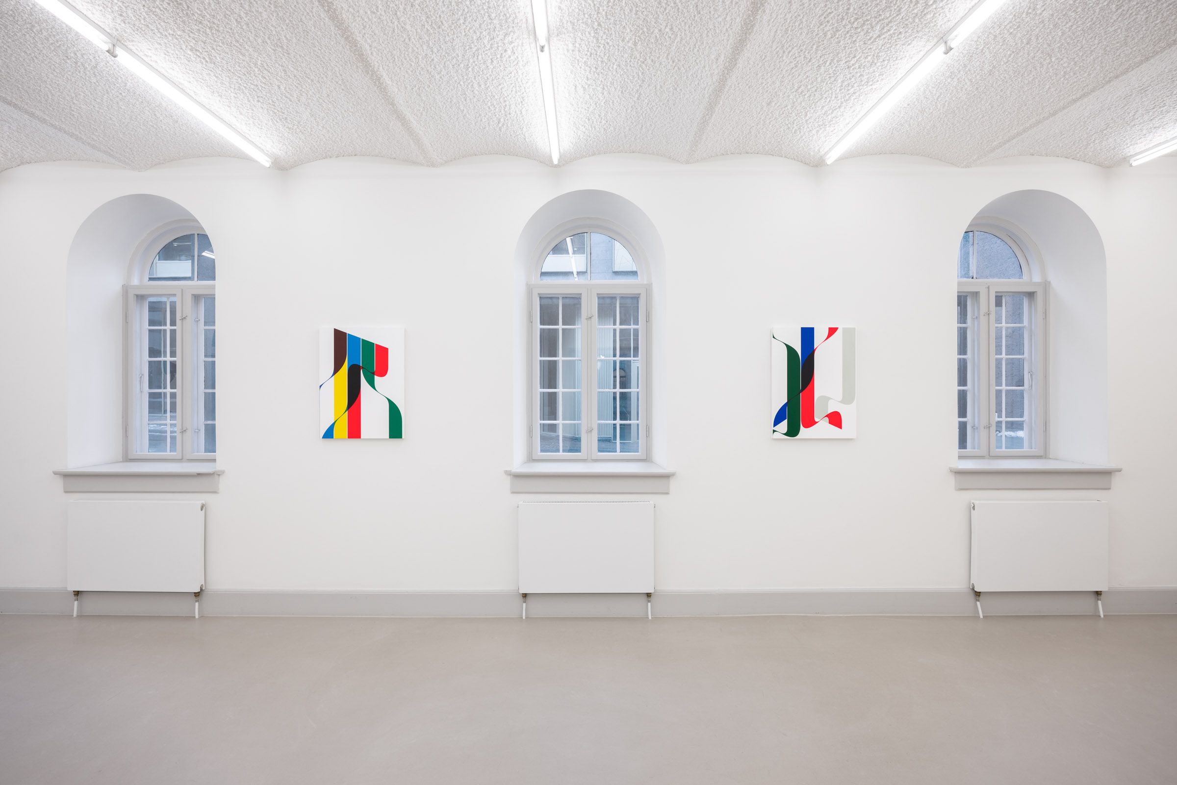 Et tomt værelse med tre vinduer, tre farverige malerier og tre hvide radiatorer på hver side af vinduer.