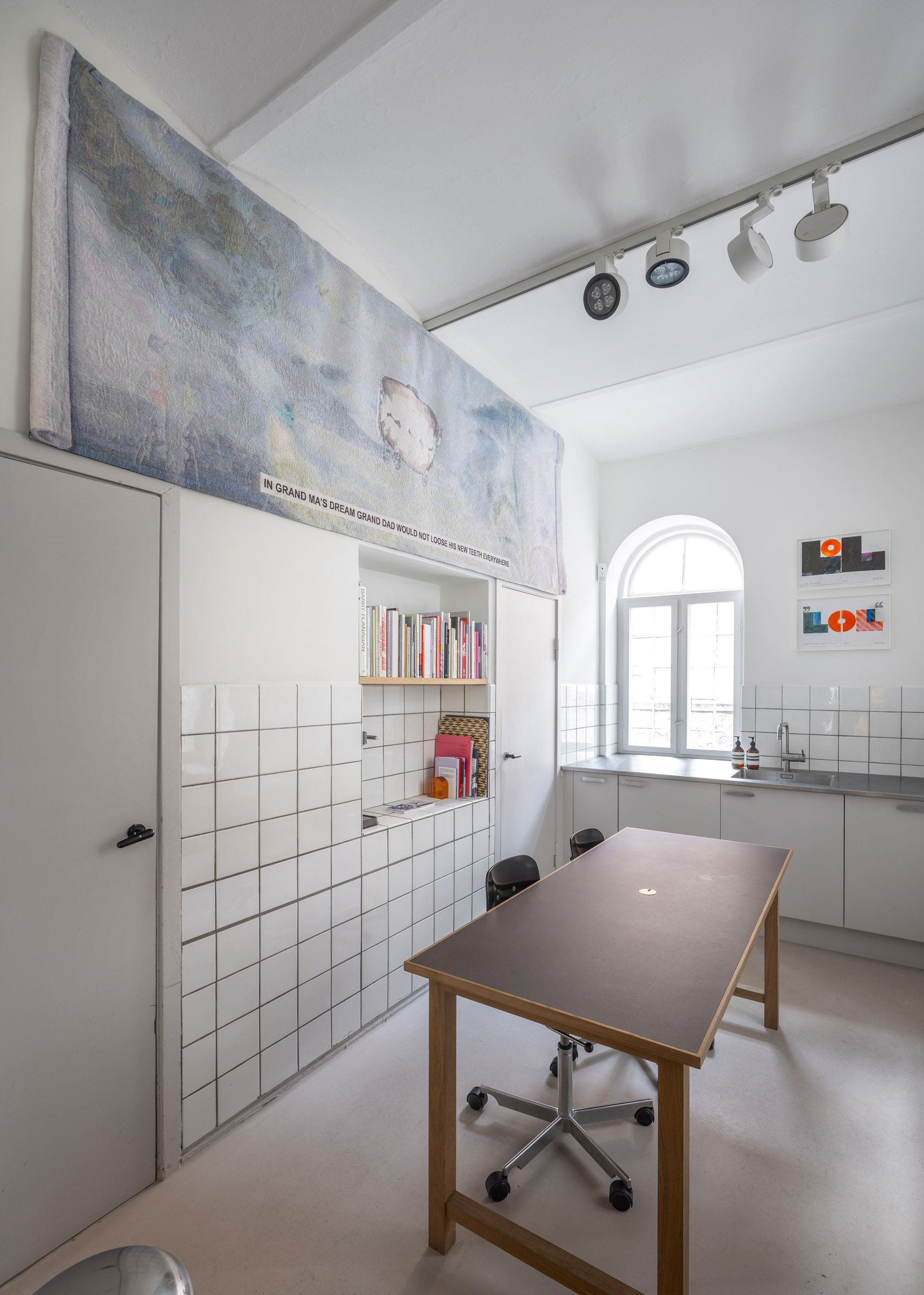 Indenfor et minimalistisk rum med hvide vægge, et bord og en stol samt højrekredsede lysekroner og et vinduesglas med tekst