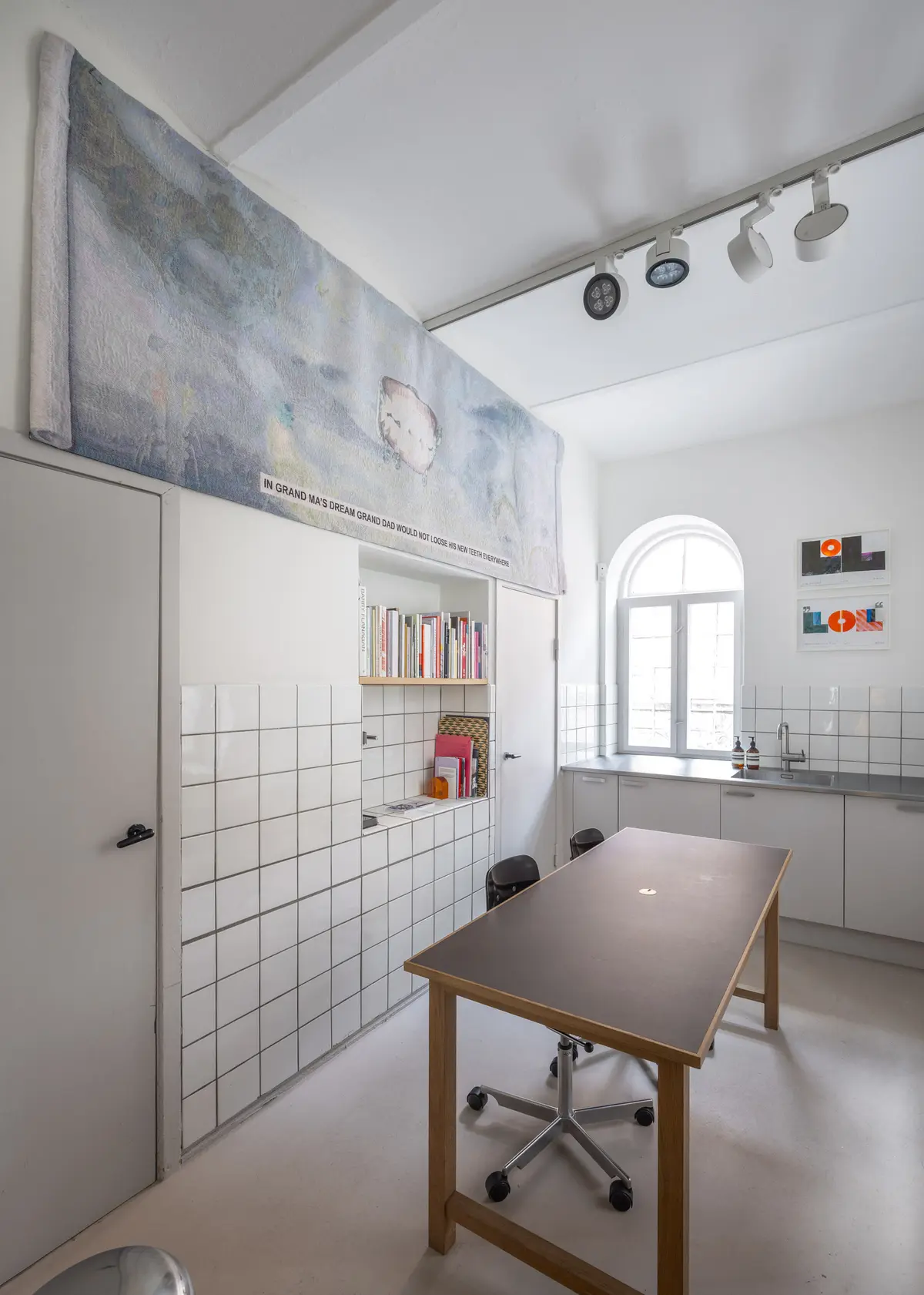 Indenfor et minimalistisk rum med hvide vægge, et bord og en stol samt højrekredsede lysekroner og et vinduesglas med tekst