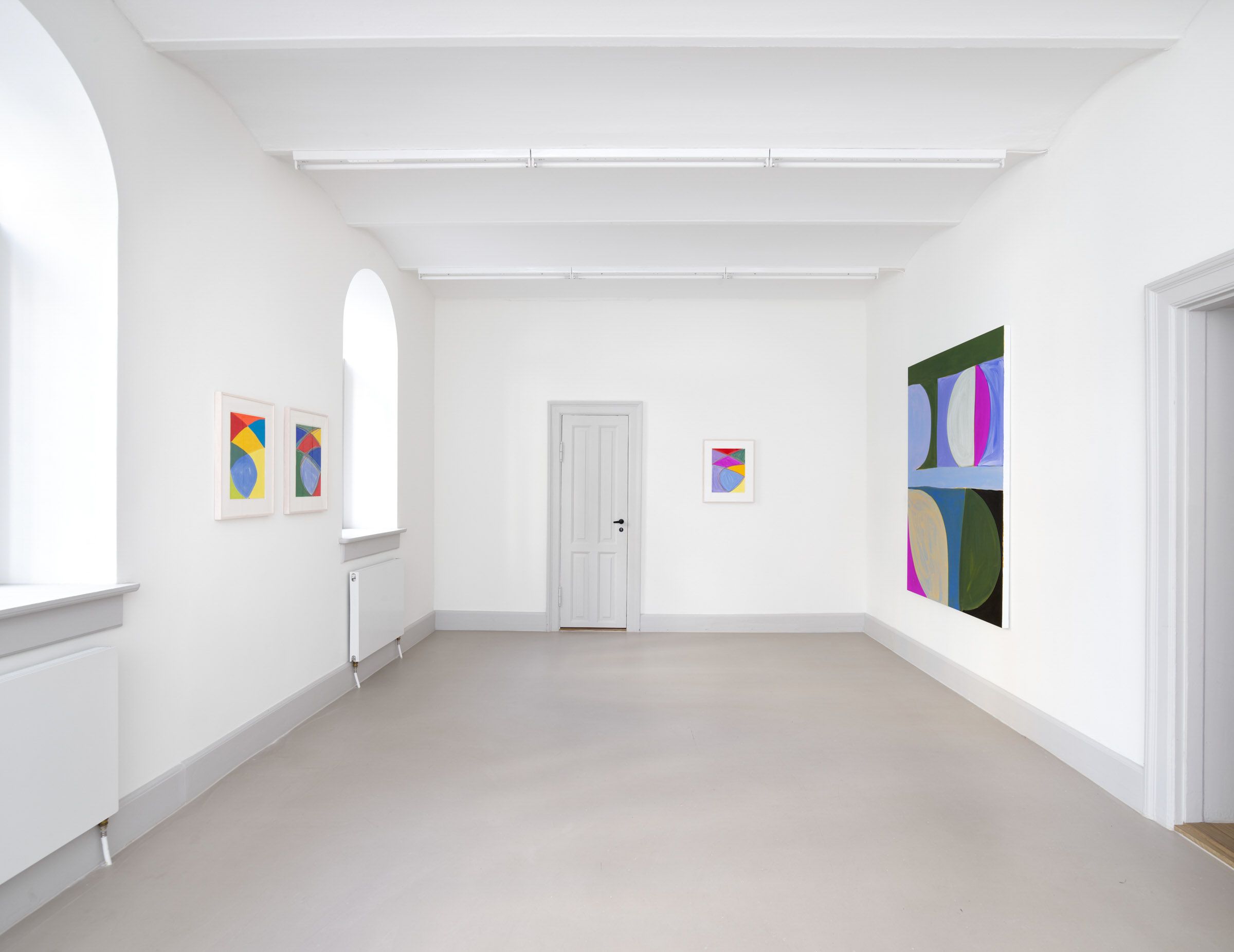 En hvid væg med tre malerier og et hvidt dør. Der er lysekroner og radiatorer. Der er en åbning med et vindue. Gulvet er