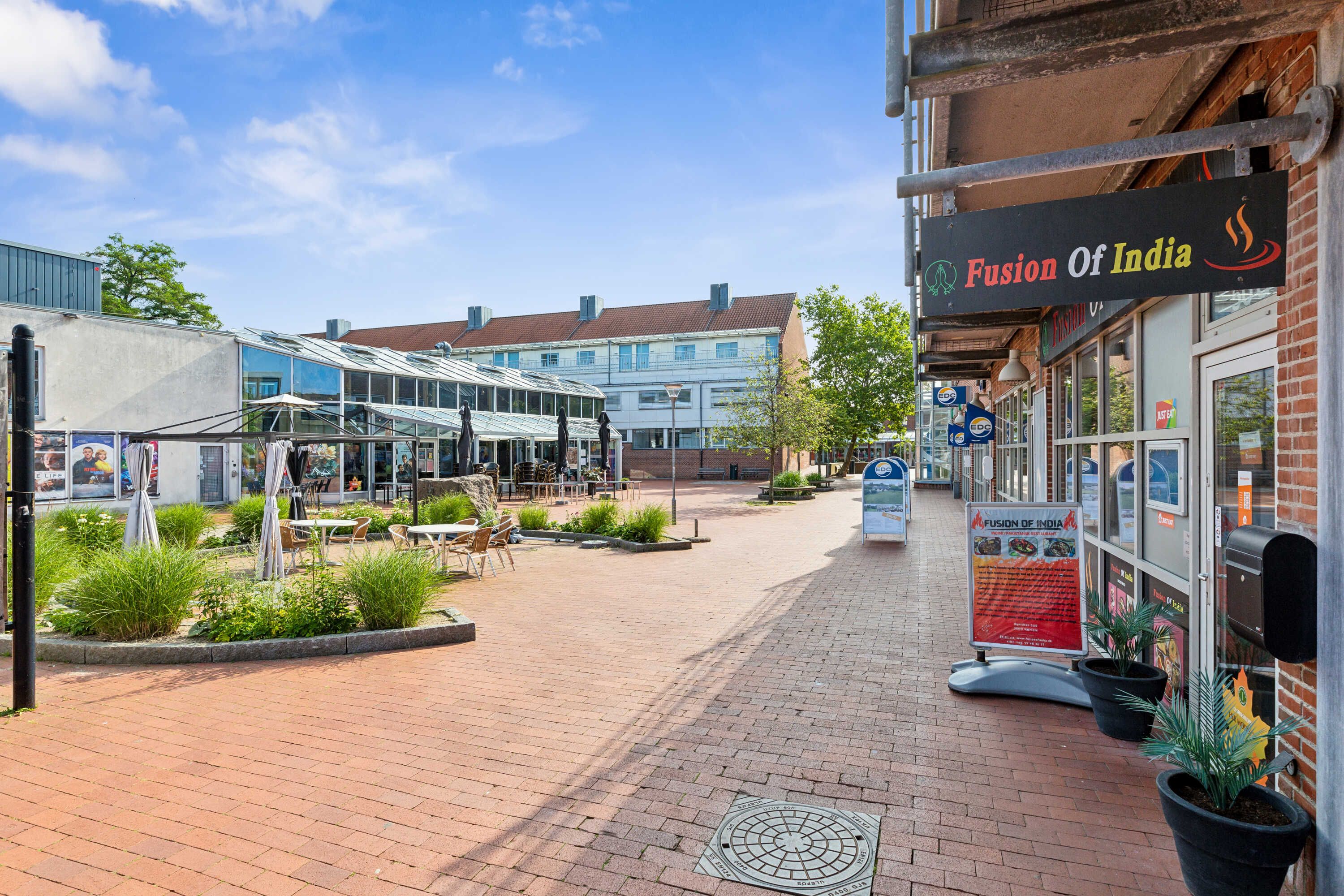 Fusion of India restaurant med grønne planter, bord og stole udenfor og en solrig dag med lyseregn i baggrunden