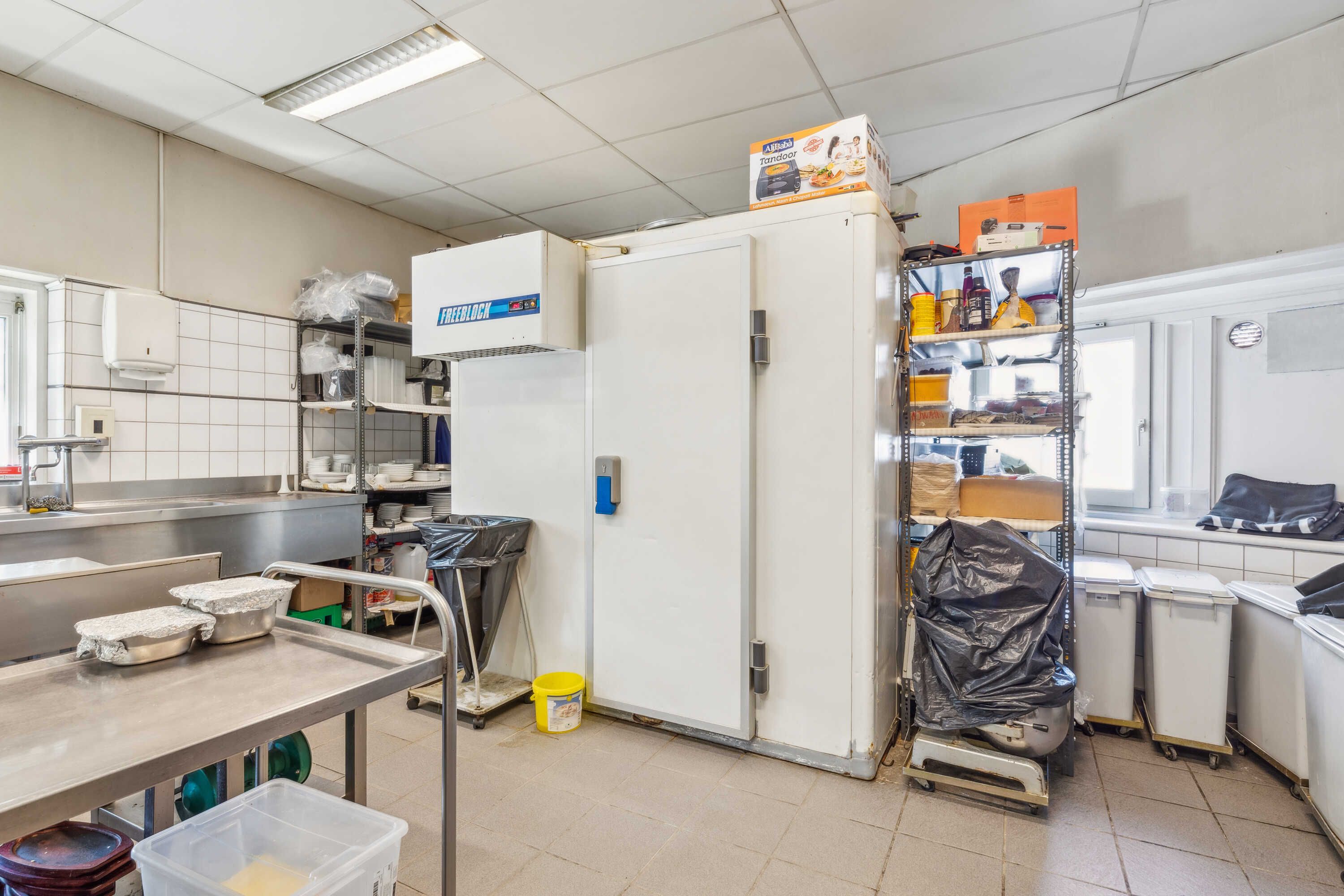 Et køkken med en stor kasse med flere ting, en frigiver, en stålbord, en bøtte og en stak plastbeholdere samt en gul spand