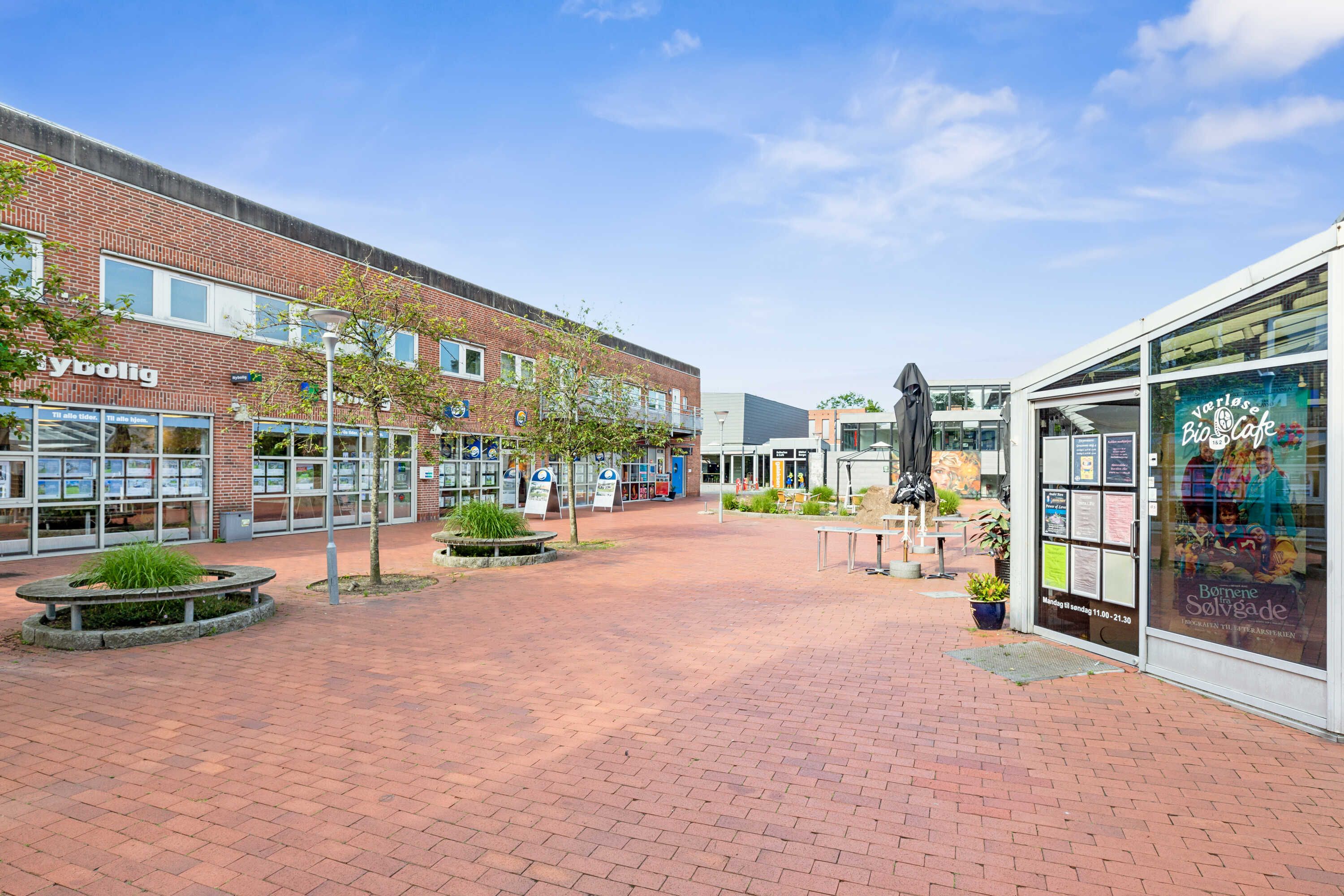 Gade med tomme gavl og træer og plante i midten, gul og rød murstensgulv, glasdøre med reklameskilte, solsenge og