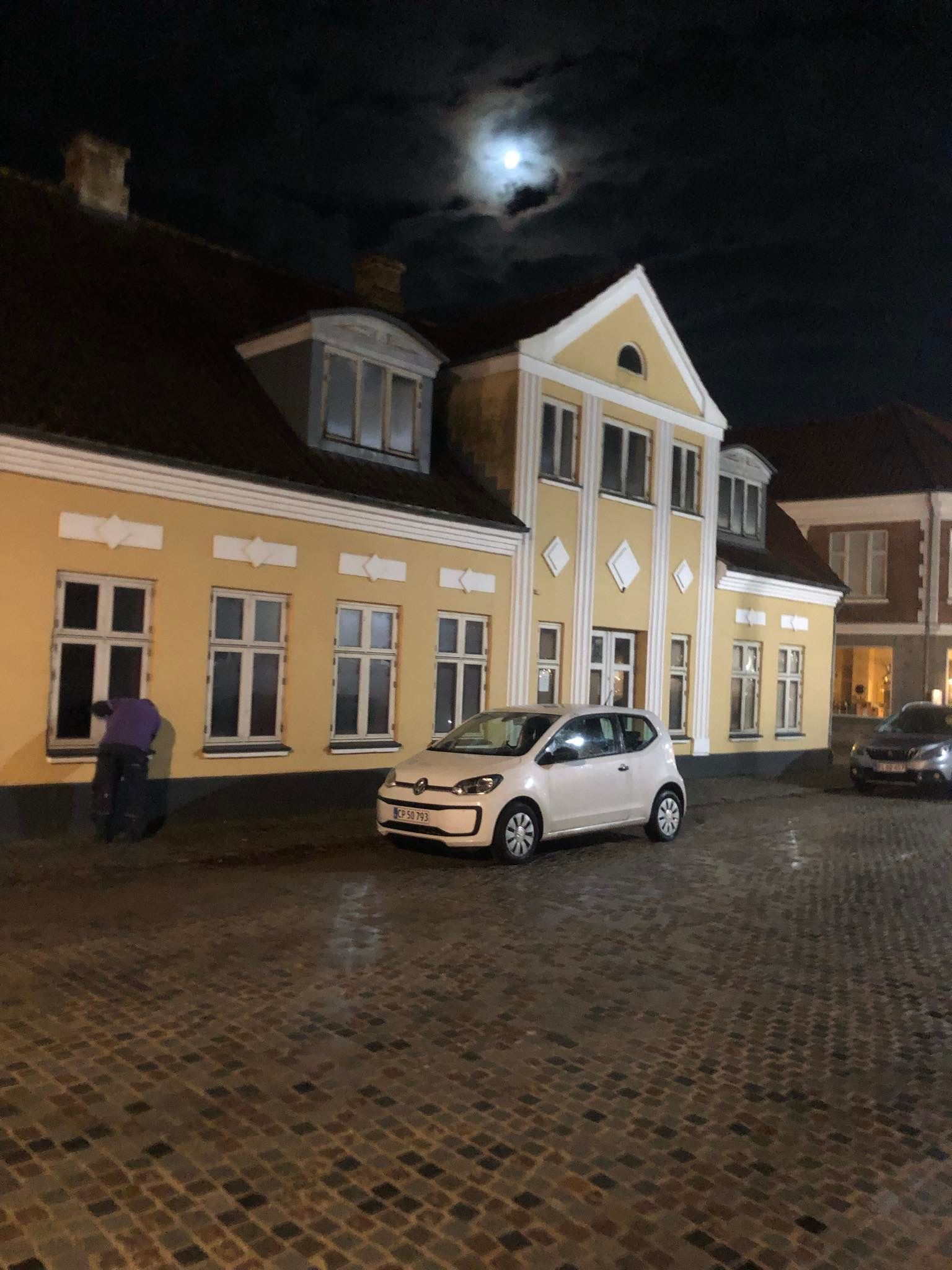 En vognparkering med en hvid Volkswagen Up og en sort bil parkeret. En mand står og ser på den hvide bil. Nattehimlen er