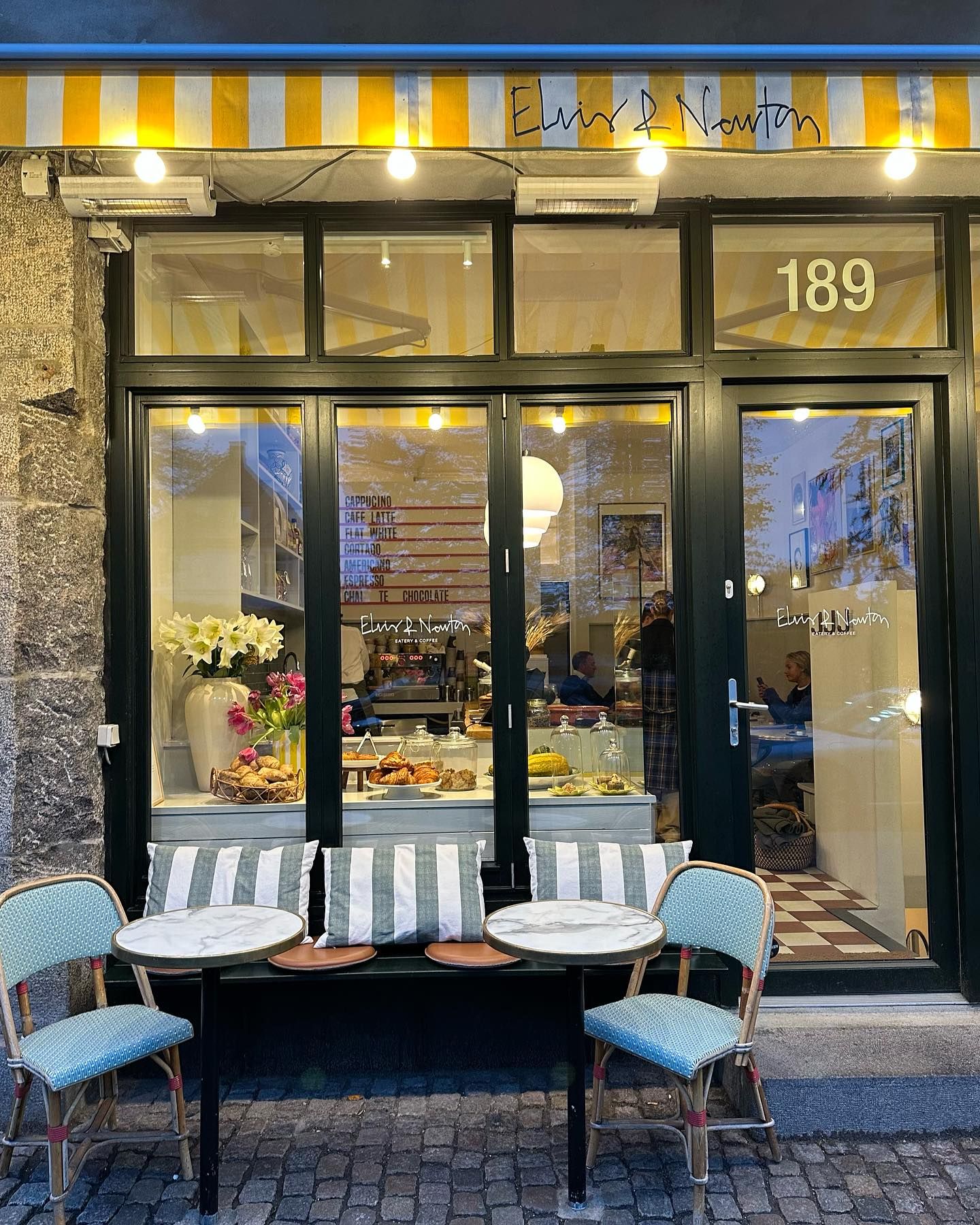 Café med bord og stole med blomster og tæpper, vinduer med udsigt til indretningen og menukort