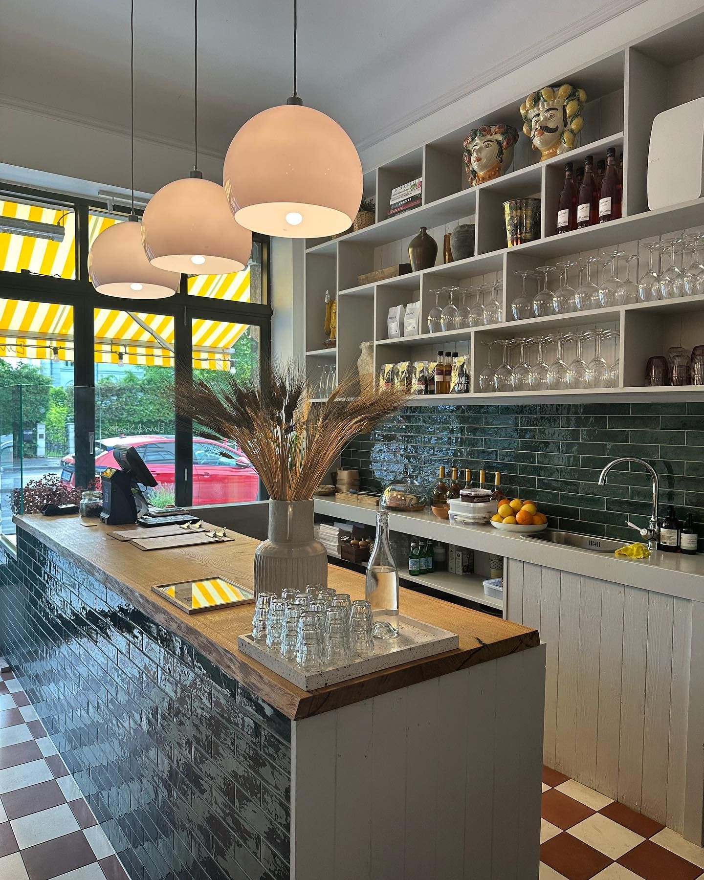 Indkøbsbord med drikkeglas og vase med hvede, hylder med flasker og glas og trægulv i caféen