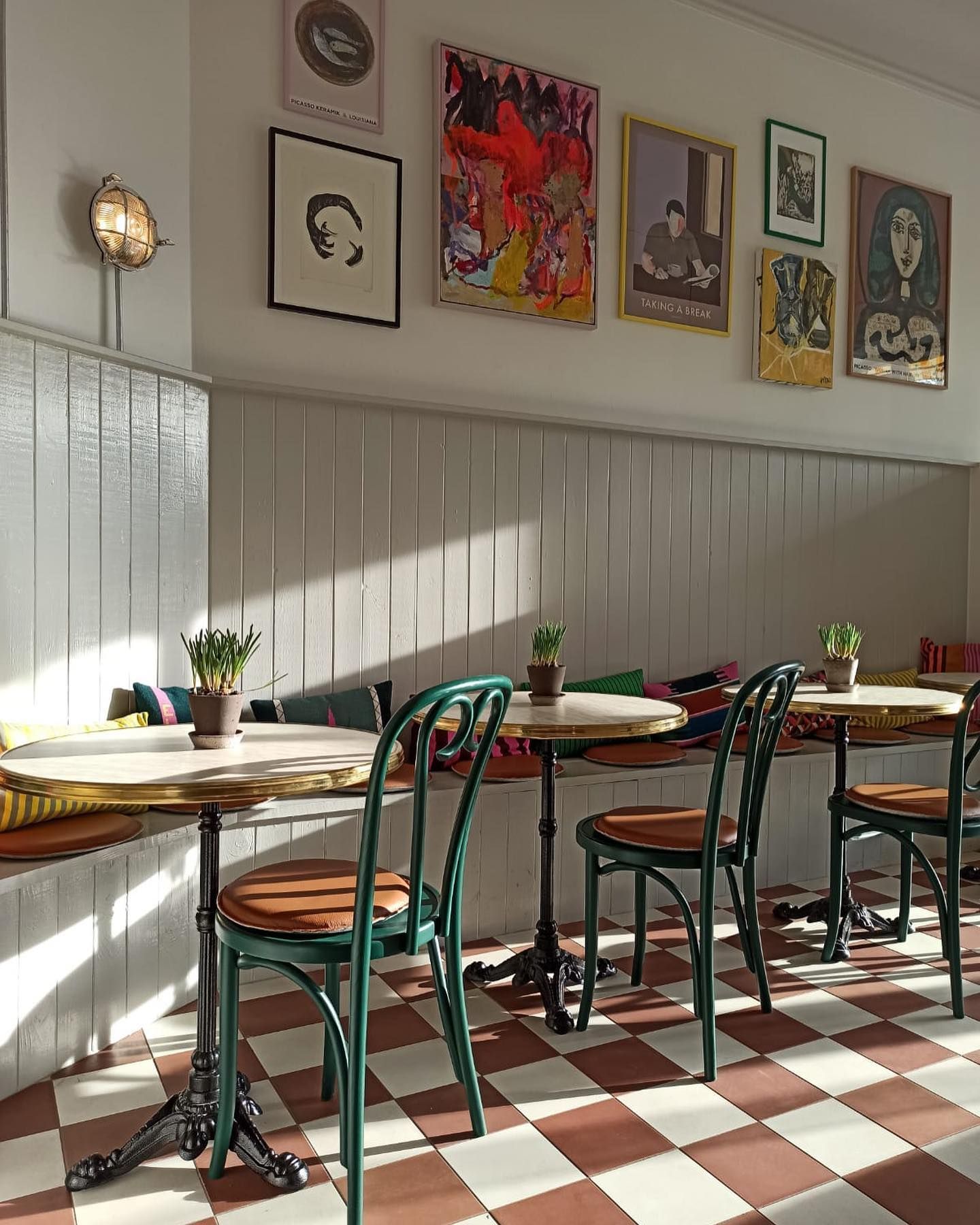En lille café med gulvbelægning i mønstret, fire små borde med grønne stole og lyserøde pude og kunstværker på væggen