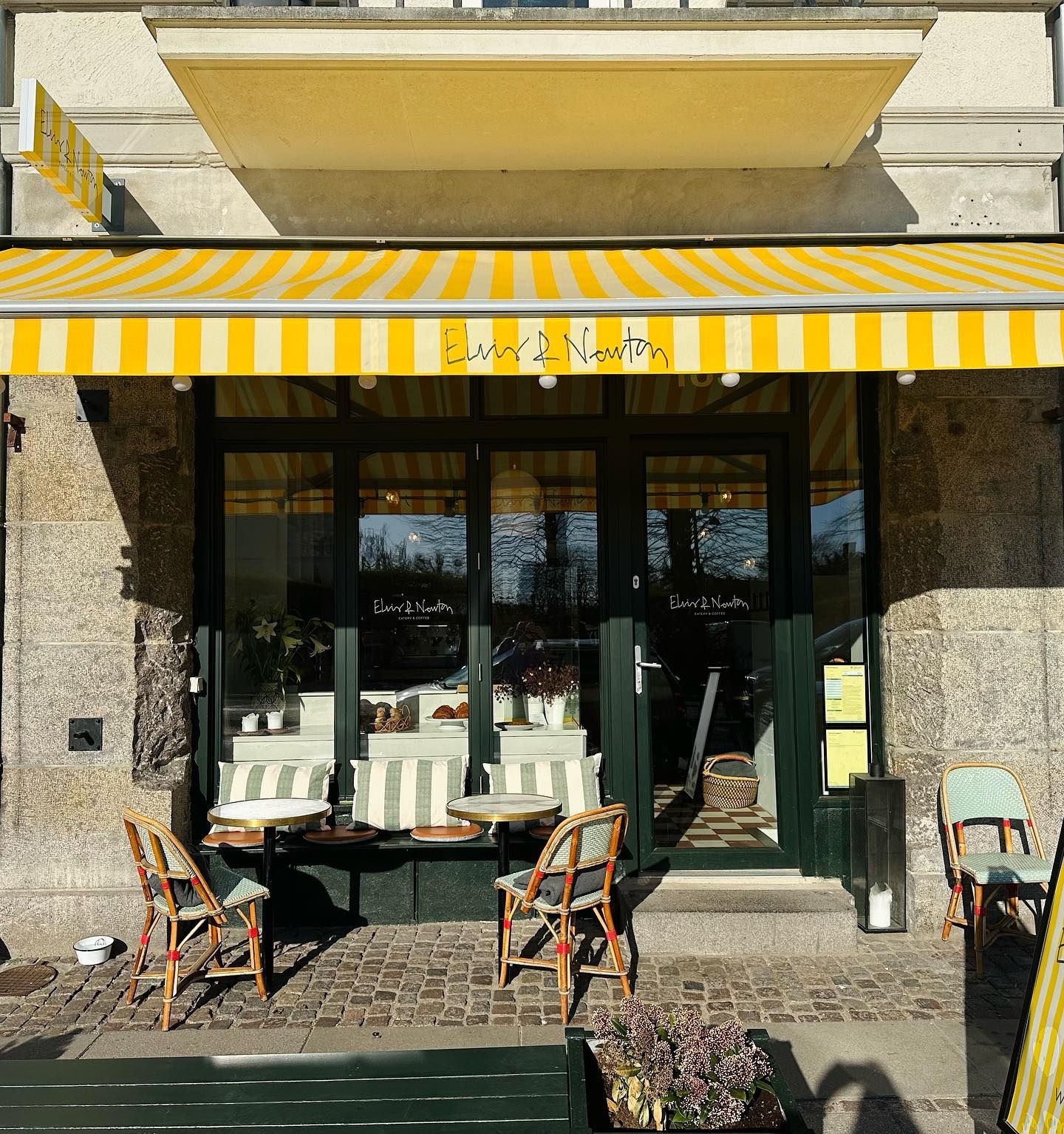 En elegant café med en gule og hvide markise, smukt møbleret med borde og stole udenfor foran bygningen