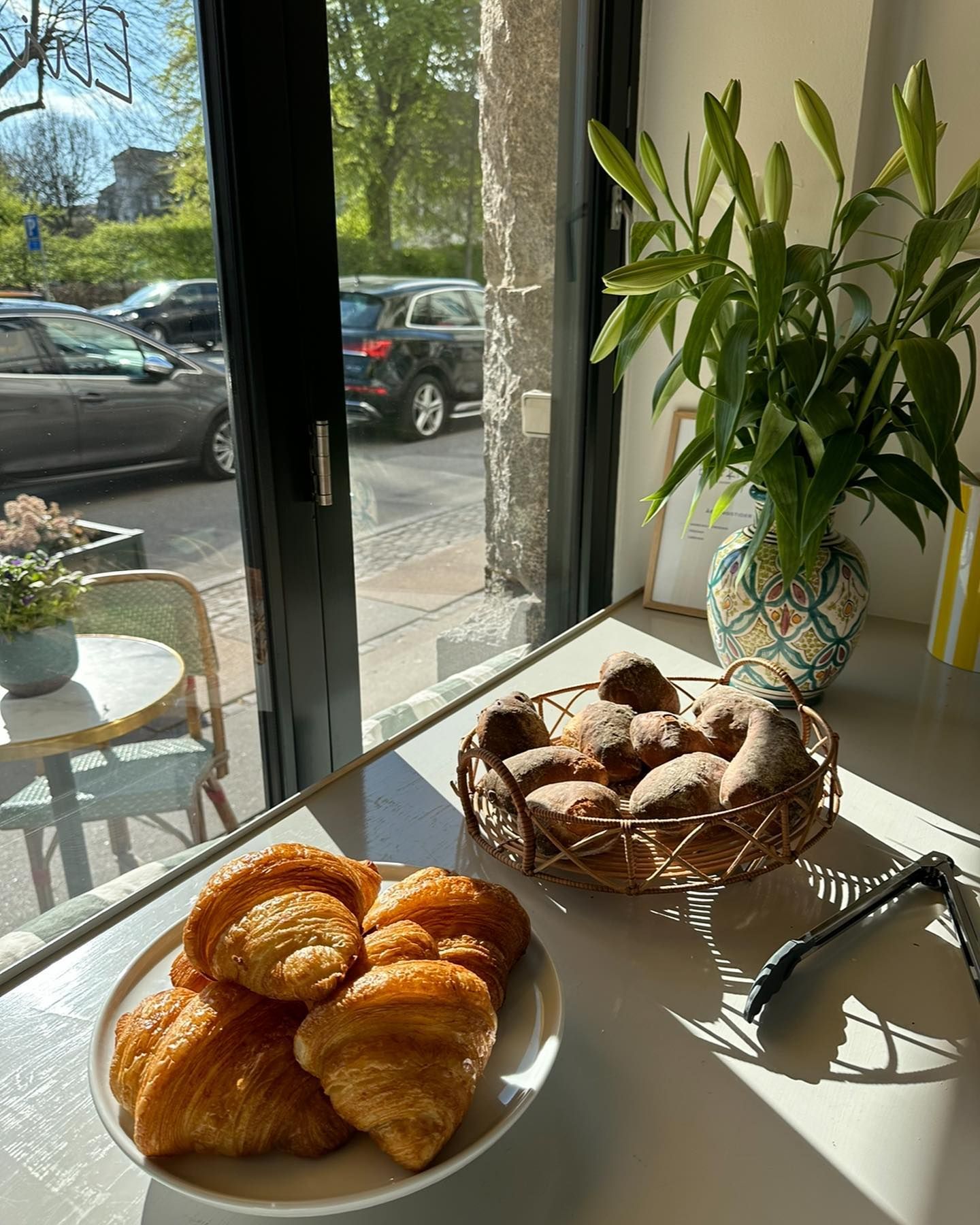 En hvid plade med flere croissanter og en kurv med bagværk på en hvid bordplade, med en vase med grønne planter og en stol