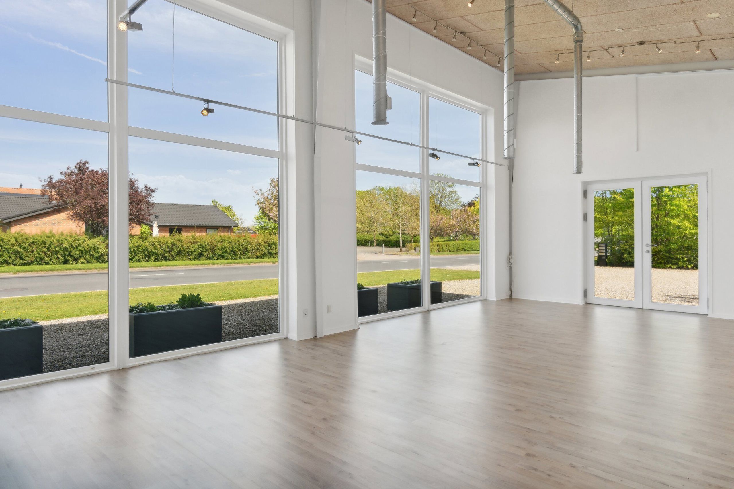 En stor, lys og rummelig lokale med lysegul gulvbelægning og store glasvinduer med udsigt til grønne planter og en landevej
