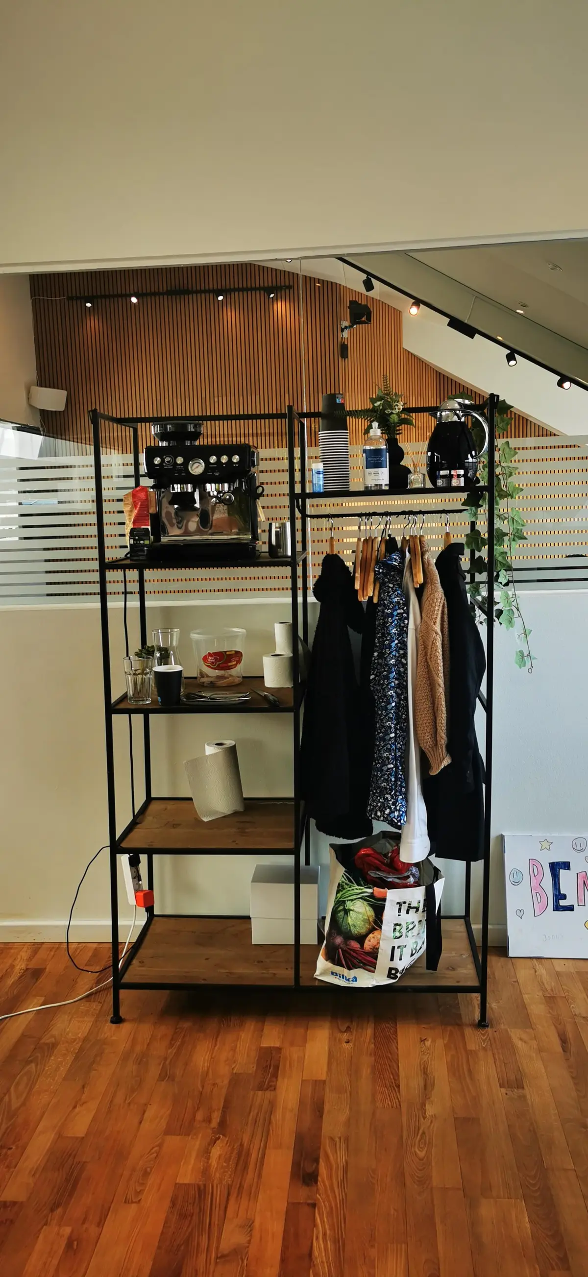 et hyggeligt kontor med en kaffemaskine, tomme kopper og tøj på et hyldesystem i et moderne kontor