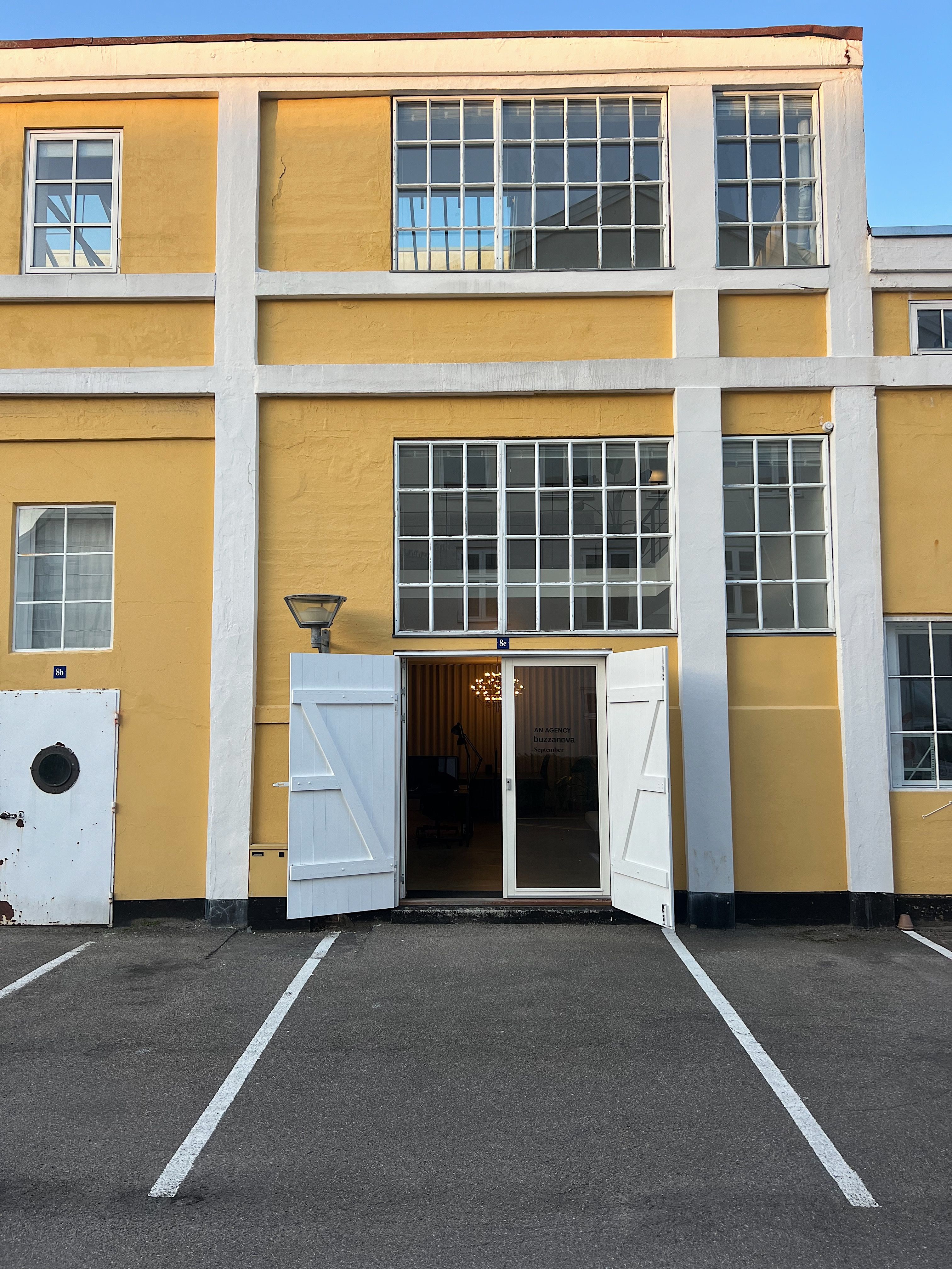 Kontorbygning med gule vægge, åbne hvide døre, glasvinduer, lysekroner og to parkeringspladser