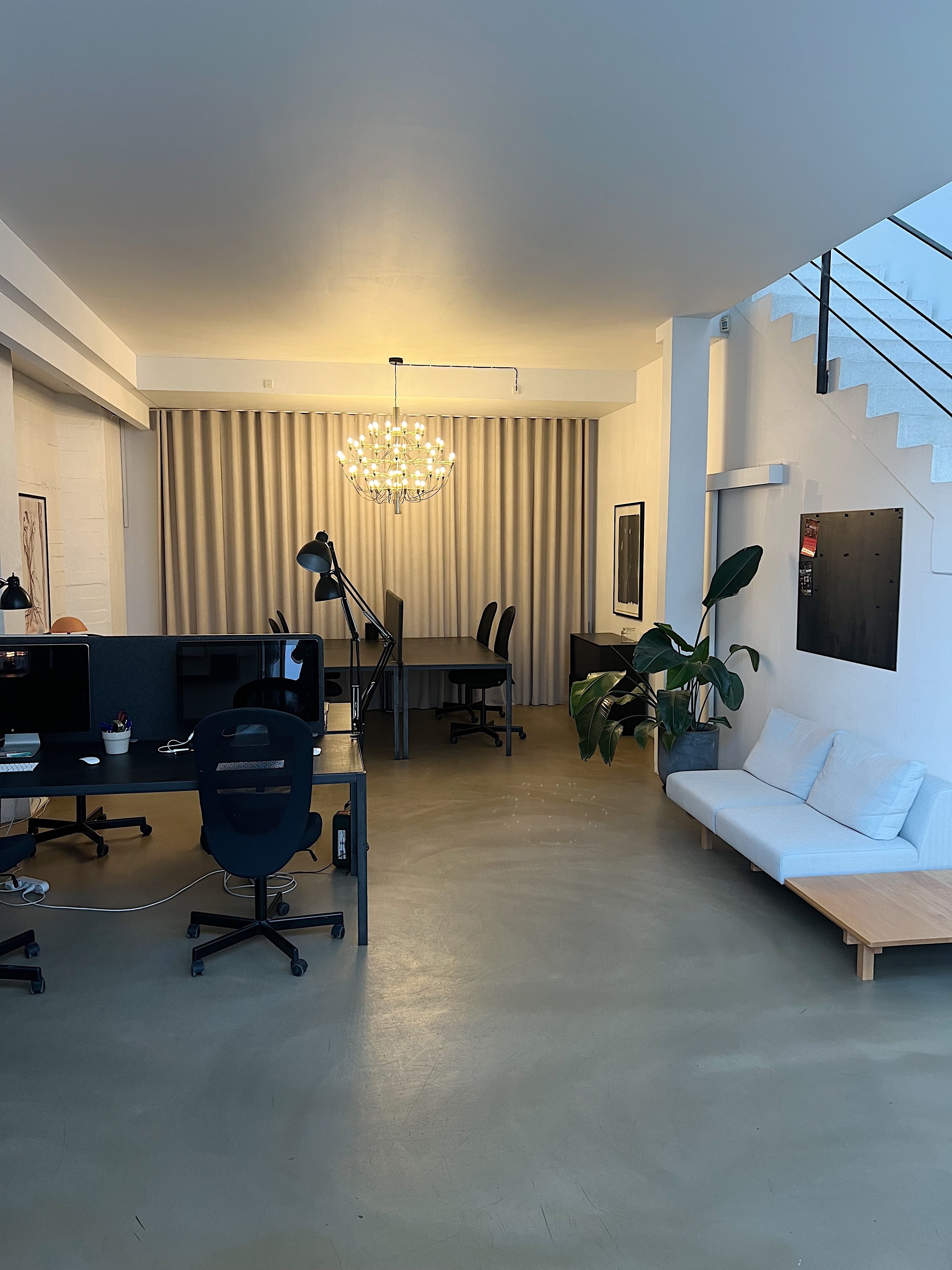 Kontor med sofa, stole, computere, lysekroner og et skrivebord med to skærme og en plante