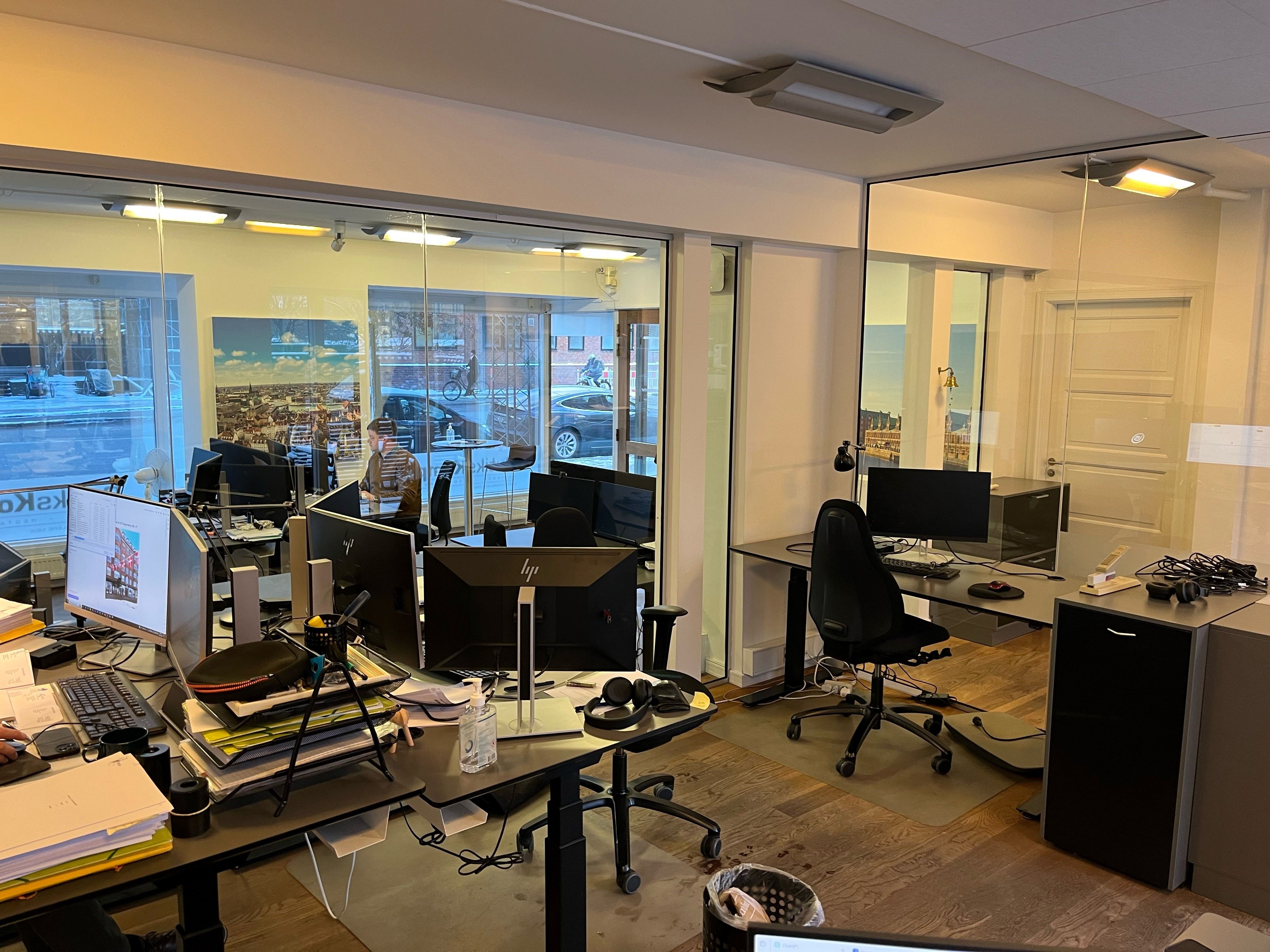 Moderne kontor med skriveborde, skærme og stole samt glasvinduer med udsigt til gade og biler udenfor
