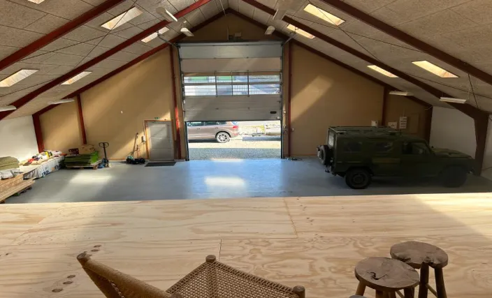 Garage med åben dør og lukket militærkøretøj, trægulve og flere træstole i lyse farver