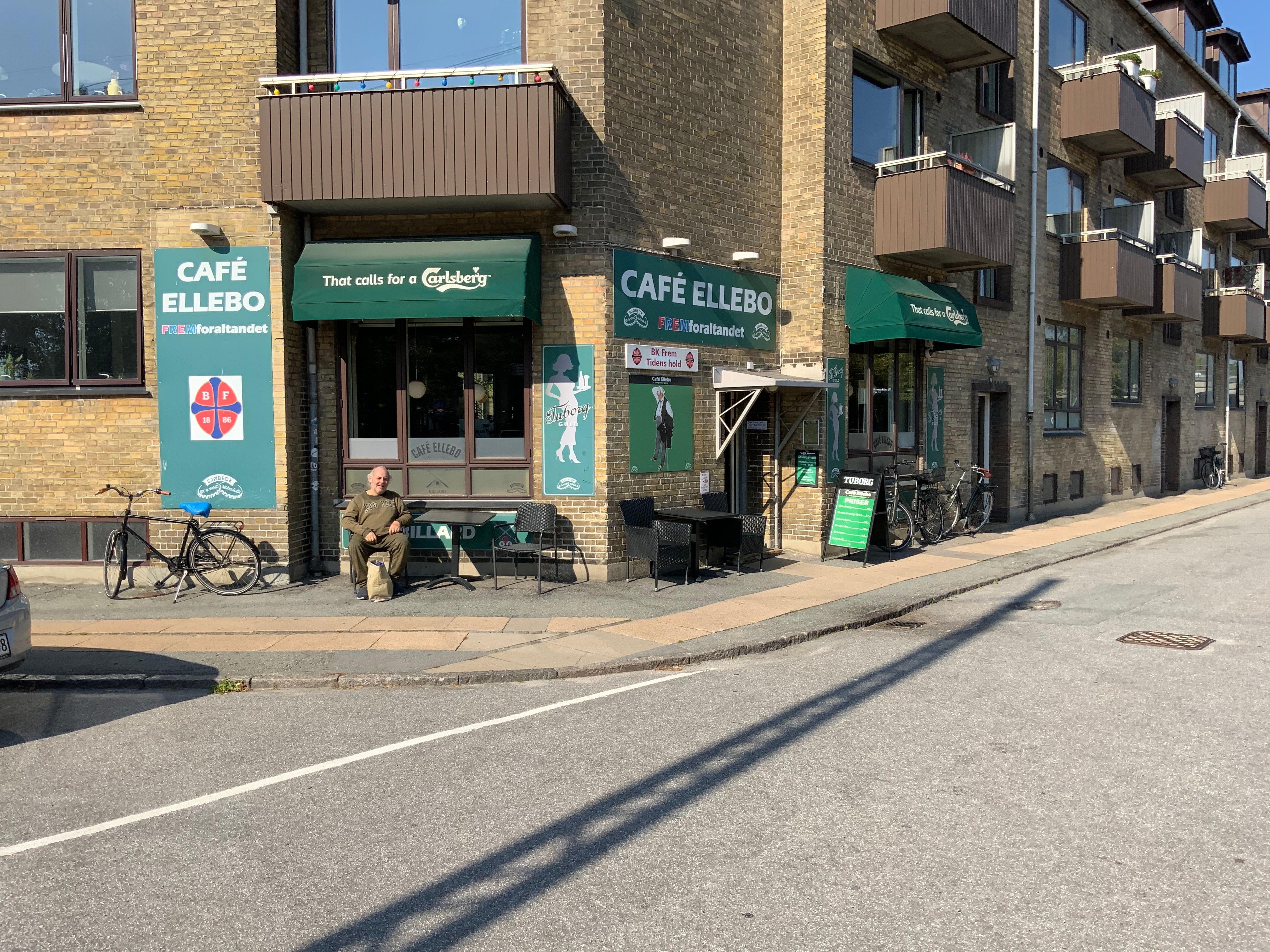 Man sidder udenfor caféen med navnet Café Ellebo. Der er stole og et bord udenfor. Der er også en cykel stillet op.