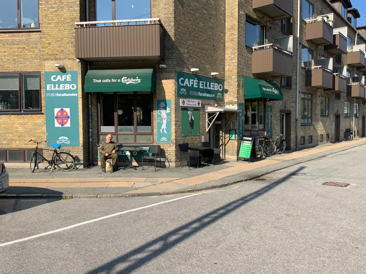 Man sidder udenfor caféen med navnet Café Ellebo. Der er stole og et bord udenfor. Der er også en cykel stillet op.