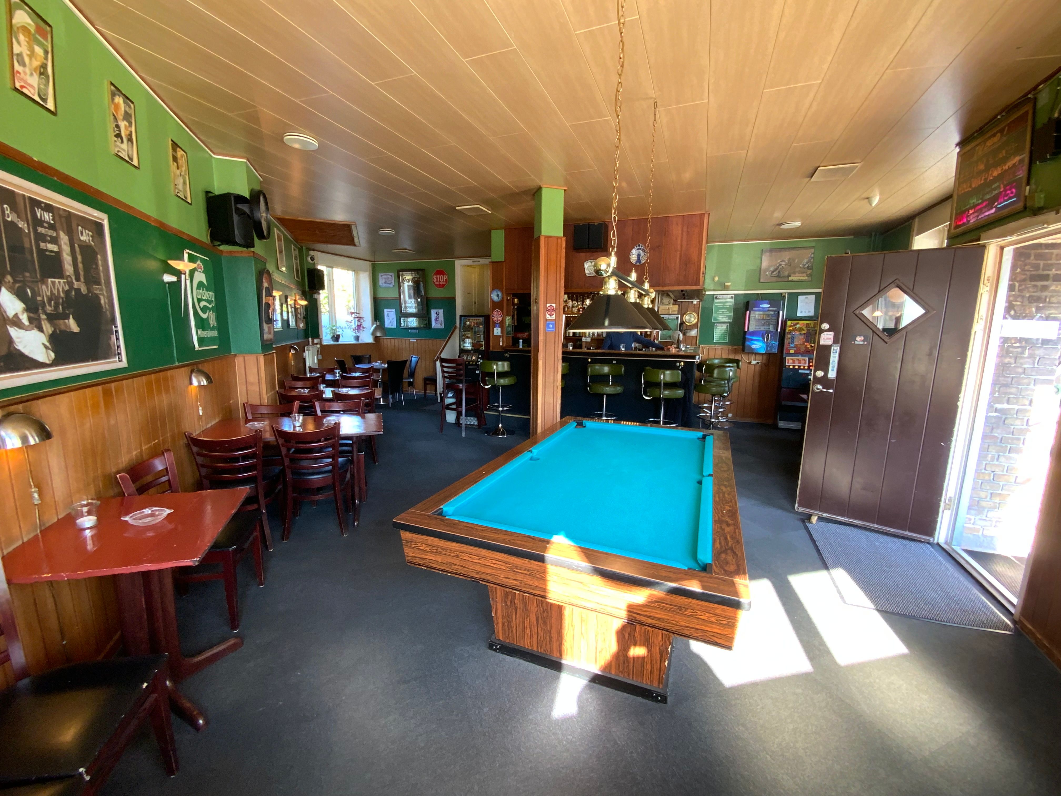 Spillebord i kafe med lyse træstole og grønne vægge. Billeder og gamle skilte hænger på væggen, og der er et smilbart ansigt